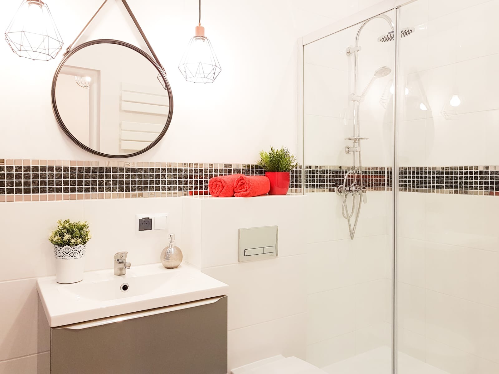 Nowoczesna łazienka z kabiną prysznicową, umywalką z szafką i okrągłym lustrem. Dekoracyjne mozaiki i oświetlenie dodają elegancji. Widoczne akcenty kolorystyczne.