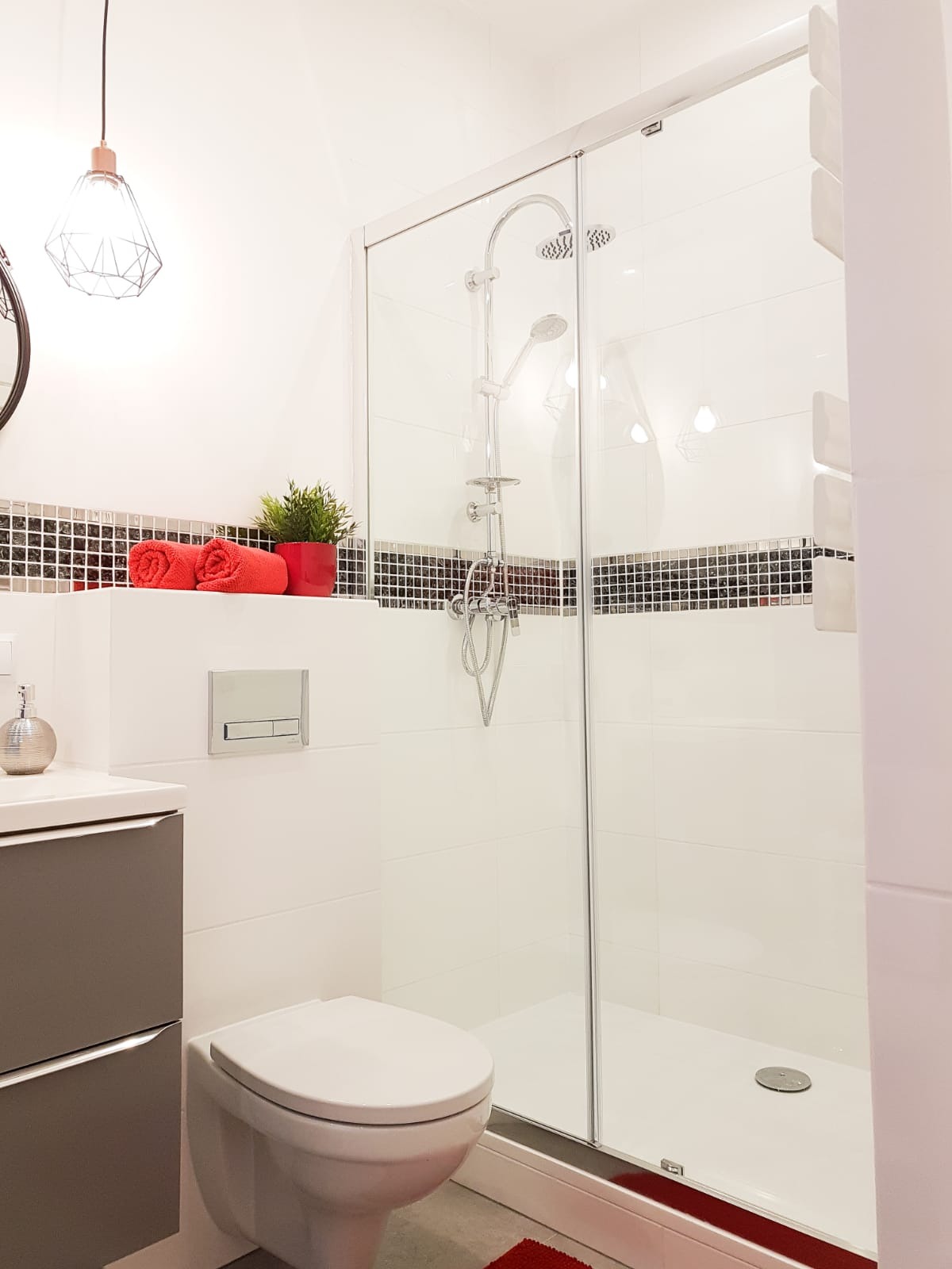 Nowoczesna łazienka z kabiną prysznicową, białe płytki z mozaiką, szare szafki pod umywalką, czerwone akcenty (ręczniki, doniczka). Geometryczna lampa wisząca.