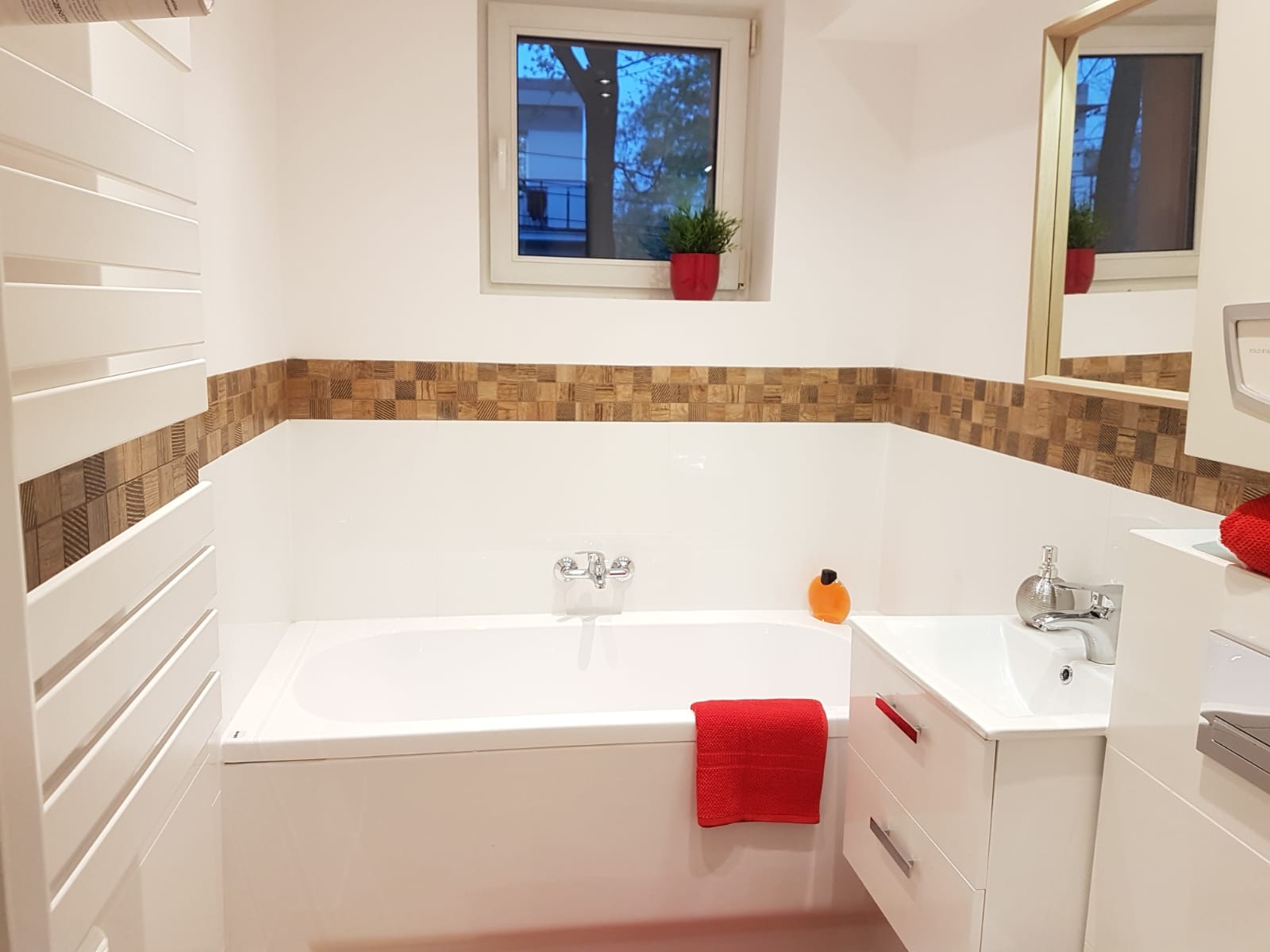 Jasna łazienka z wanną, umywalką i grzejnikiem drabinkowym. Dekoracyjne płytki imitujące drewno w pasie nad wanną. Okno z widokiem na zewnątrz i roślina w doniczce.