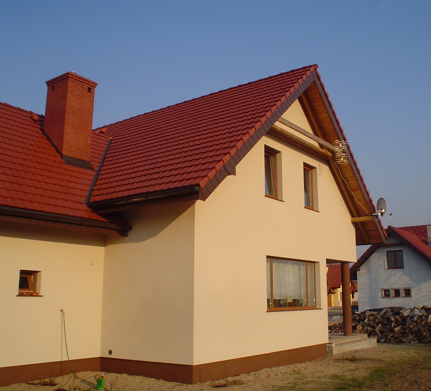 Dwukondygnacyjny dom z jasną elewacją i czerwoną dachówką, widoczne detale wykończenia, komin z cegły i antena satelitarna, w tle stos drewna.