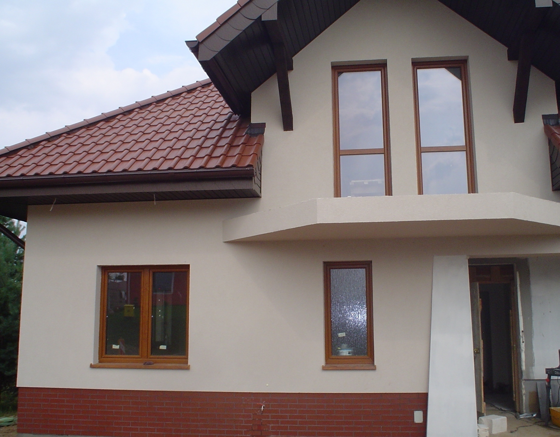 Dwukondygnacyjny dom z jasną elewacją, brązową dachówką i oknami w drewnianych ramach. Część elewacji wykończona cegłą klinkierową. Widoczne elementy rusztowania i materiały budowlane przy wejściu.