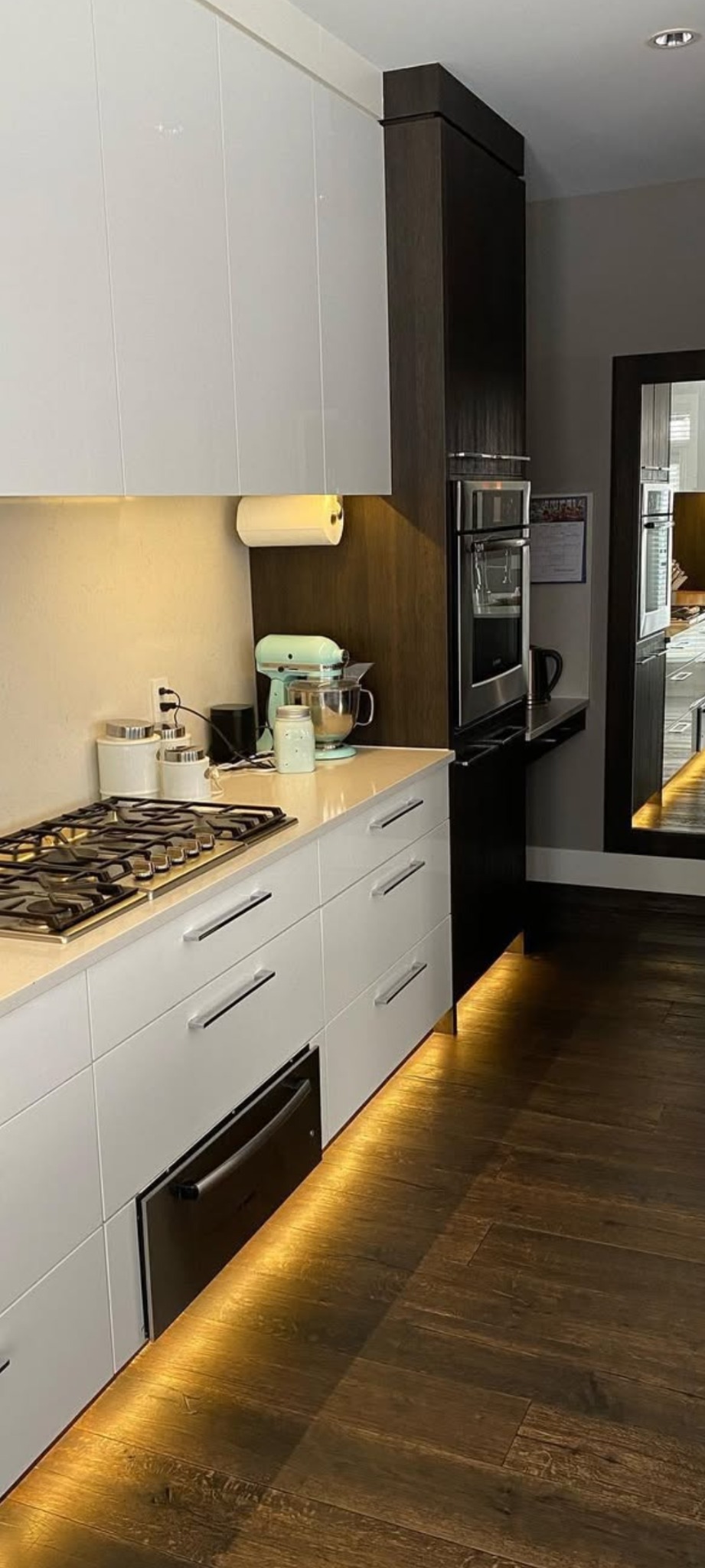 Nowoczesna kuchnia z białymi szafkami, drewnianymi akcentami i podświetleniem LED. Na blacie mikser, a w tle widoczne odbicie w lustrze.