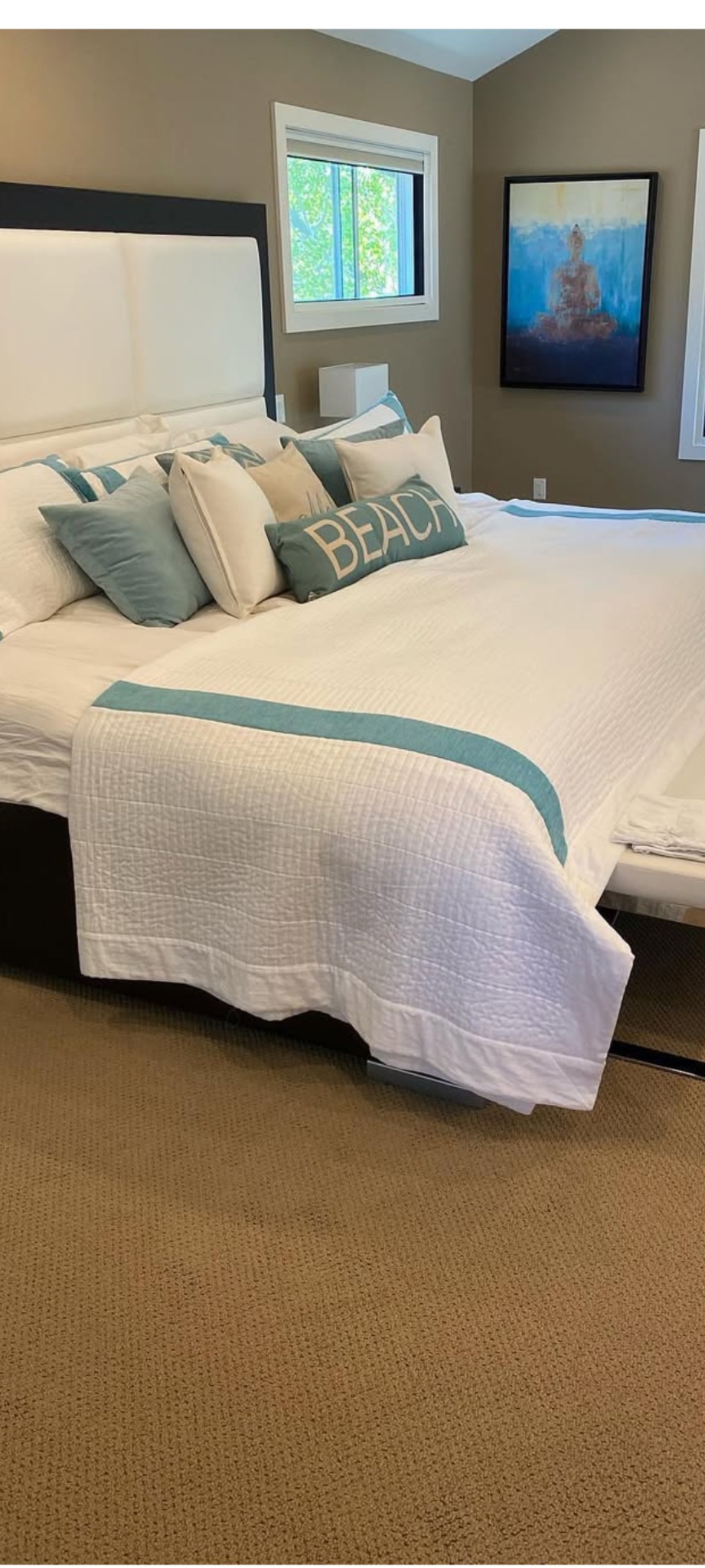 Wnętrze sypialni z dużym łóżkiem, białym obrusem z turkusowymi akcentami, poduszkami i obrazem na ścianie w stonowanych kolorach ziemi.