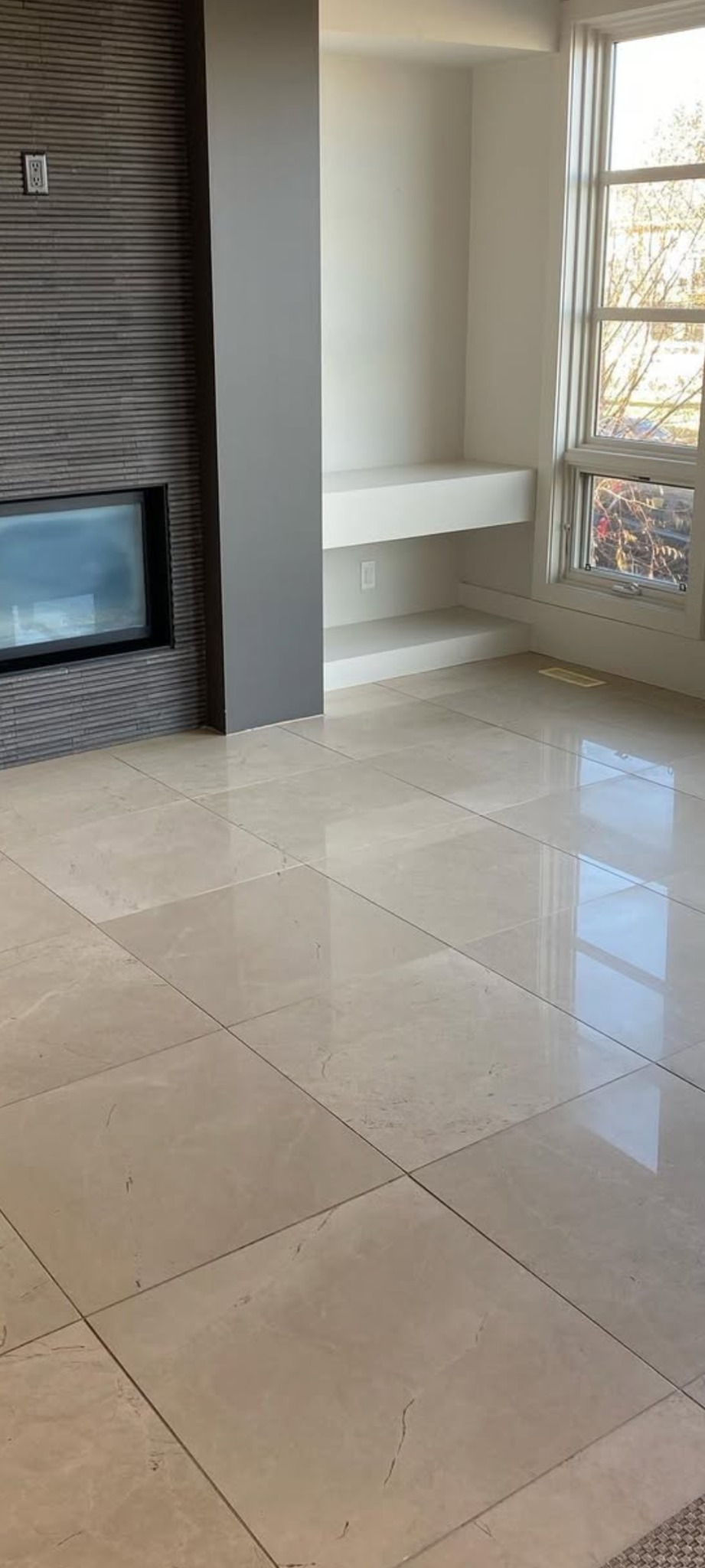 Wnętrze pokoju z marmurową podłogą, kominkiem z szarej cegły i wnęką z półkami. Widoczne okno z widokiem na drzewa. Jasne, przestronne pomieszczenie.