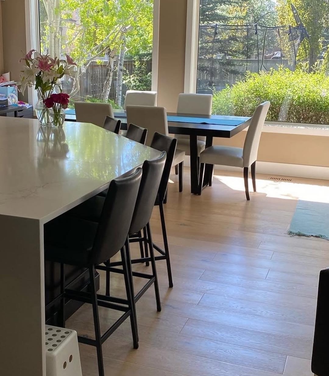 Jasne wnętrze jadalni z wyspą kuchenną i widokiem na zieleń za oknem. Kwiaty w wazonie na blacie. Krzesła barowe przy wyspie, stół z krzesłami w tle.