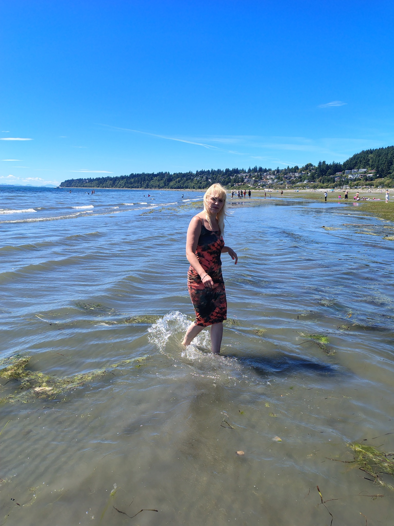 Kobieta w sukience brodzi w płytkiej wodzie morskiej, z widoczną linią brzegową i grupą ludzi w tle, w słoneczny dzień. Woda lekko wzburzona, z wodorostami.