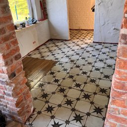 Remontivo - Układanie wzorzystej terakoty w pokoju z fragmentami ceglanej ściany. Część podłogi wyłożona drewnopodobnymi płytkami. Widoczne narzędzia i poziomica.