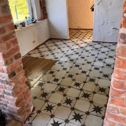 Układanie wzorzystej terakoty w pokoju z fragmentami ceglanej ściany. Część podłogi wyłożona drewnopodobnymi płytkami. Widoczne narzędzia i poziomica.