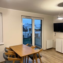 Remontivo - Jasne wnętrze pokoju z drewnianym stołem i krzesłami, widokiem na osiedle przez okno. Minimalistyczny design, parkiet i nowoczesne oświetlenie.