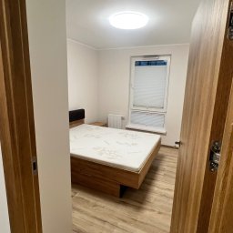 Remontivo - Jasne wnętrze sypialni z drewnianą podłogą i łóżkiem, widziane zza otwartych drzwi. Minimalistyczny wystrój, białe ściany, okno z żaluzjami. Ujęcie z perspektywy wejścia.