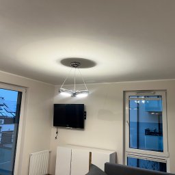 Remontivo - Jasne wnętrze pokoju z nowoczesną lampą sufitową, telewizorem na ścianie i białymi meblami. Widok na zewnątrz przez okno balkonowe.