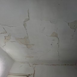 Remontivo - Popękany, biały sufit i fragment ściany w rogu pokoju, z widocznymi ubytkami i odpryskami farby, ukazujący zły stan powierzchni przed remontem.