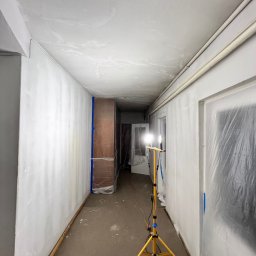 Remontivo - Korytarz w trakcie przygotowań do malowania: ściany zagruntowane, meble i drzwi zabezpieczone folią, lampa budowlana oświetla przestrzeń. Widoczny wąż odkurzacza.