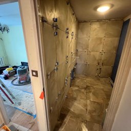 Remont łazienki: układanie płytek imitujących marmur na ścianach z użyciem systemu poziomowania. Widok z korytarza, sprzęt budowlany w tle.