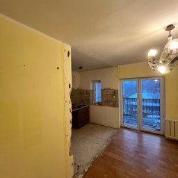 Remontivo - Remont mieszkania: widok na pokój z fragmentem kuchni w trakcie prac. Częściowo zerwane płytki, odsłonięta ściana, parkiet. Okno balkonowe z widokiem na zewnątrz.