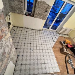 Remontivo - Układanie płytek podłogowych w trakcie remontu mieszkania. Widoczne narzędzia, płytki z wzorem i krzyżyki dystansowe. Częściowo wykończona podłoga, surowe ściany.