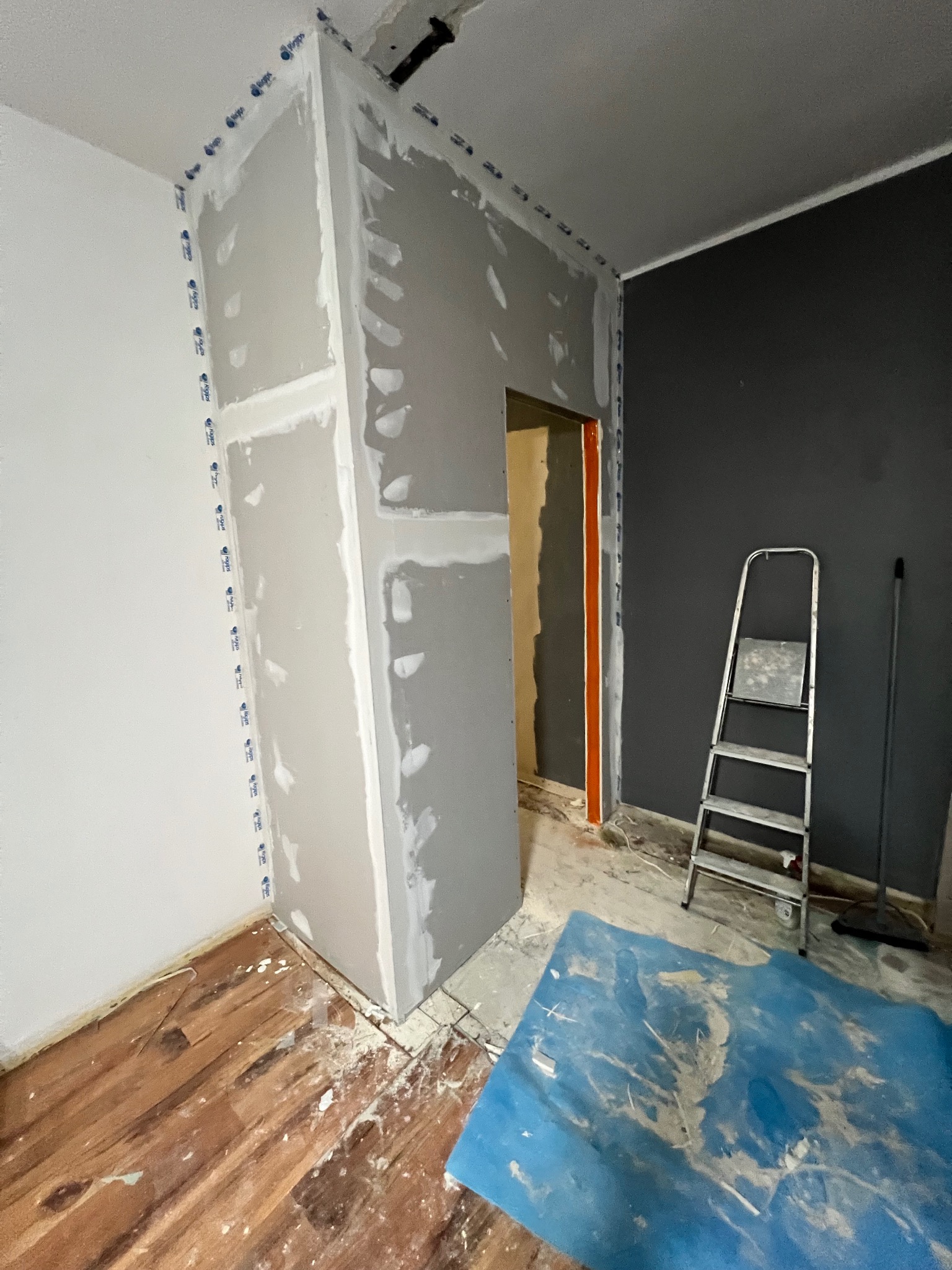 Montaż płyt gipsowo-kartonowych na ścianie z widocznymi łączeniami i taśmą zabezpieczającą. W tle drabina i pomalowana na szaro ściana.