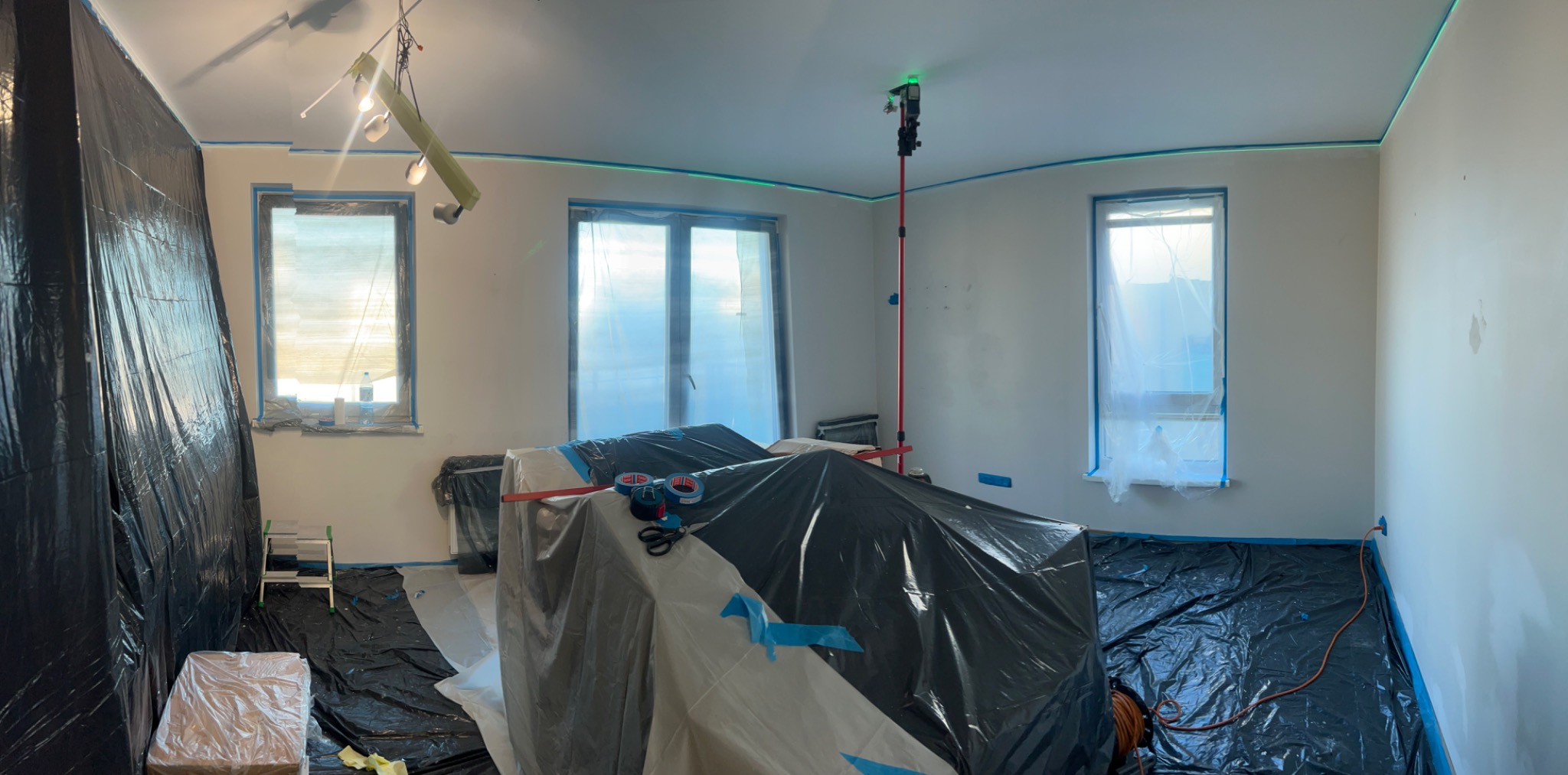 Pokój przygotowany do malowania: okna i meble zabezpieczone folią, ściany z poziomą linią lasera. Widoczne narzędzia malarskie i oświetlenie.