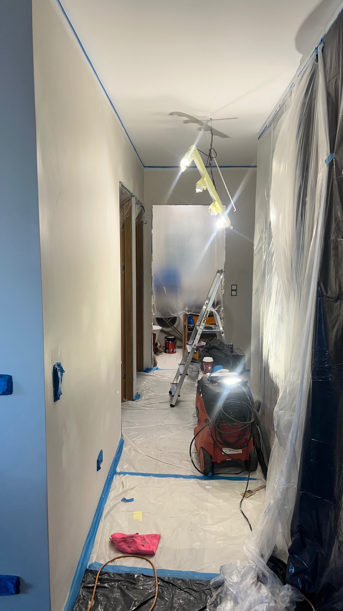 Korytarz w trakcie malowania: ściany zabezpieczone taśmą, podłoga folią ochronną, widoczna drabina i sprzęt malarski. Przygotowania do malowania ścian.