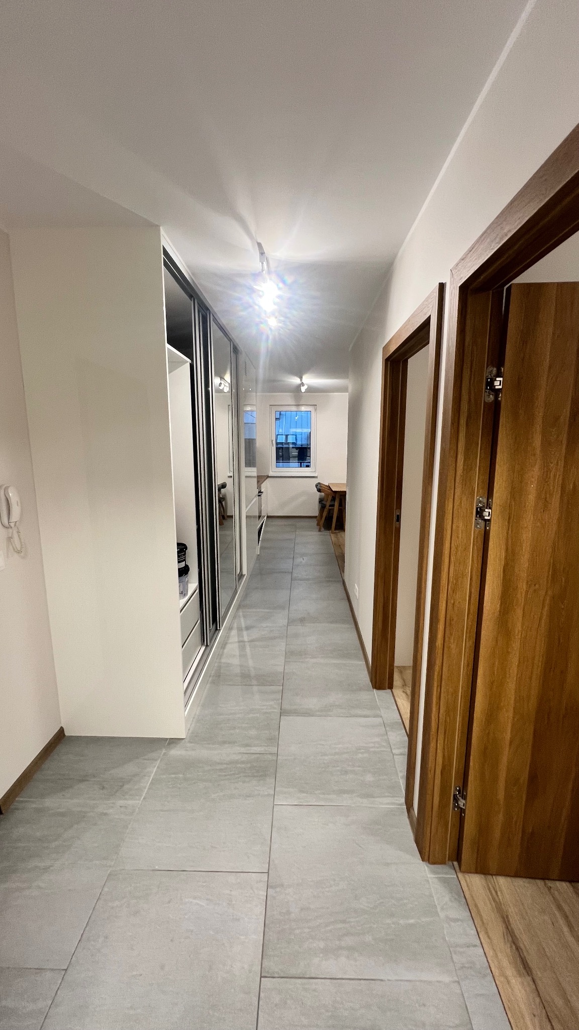 Nowoczesny korytarz z szarą podłogą, zabudowaną szafą z lustrami i drewnianymi drzwiami. Widok w głąb mieszkania, oświetlenie sufitowe.