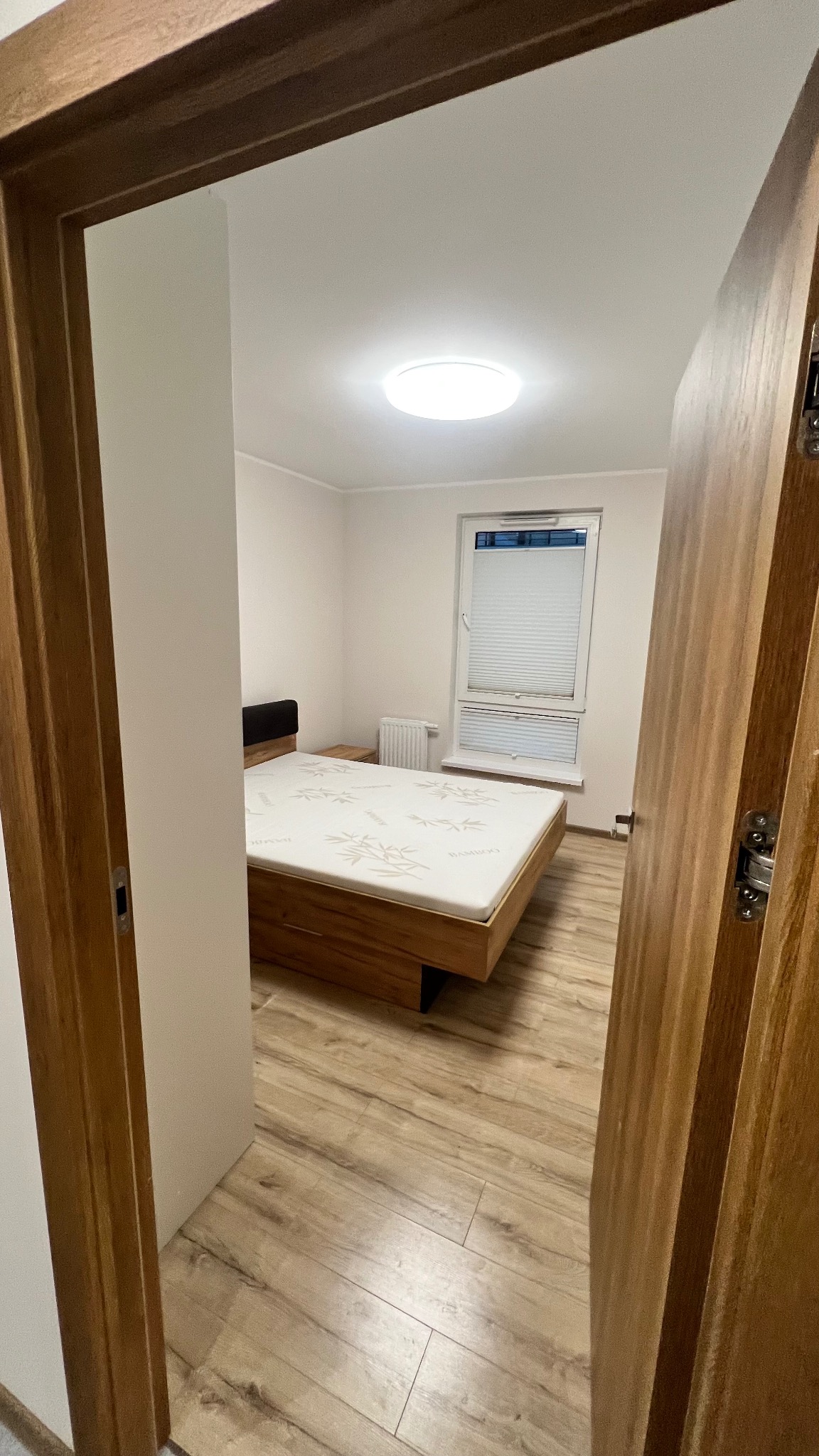 Jasne wnętrze sypialni z drewnianą podłogą i łóżkiem, widziane zza otwartych drzwi. Minimalistyczny wystrój, białe ściany, okno z żaluzjami. Ujęcie z perspektywy wejścia.