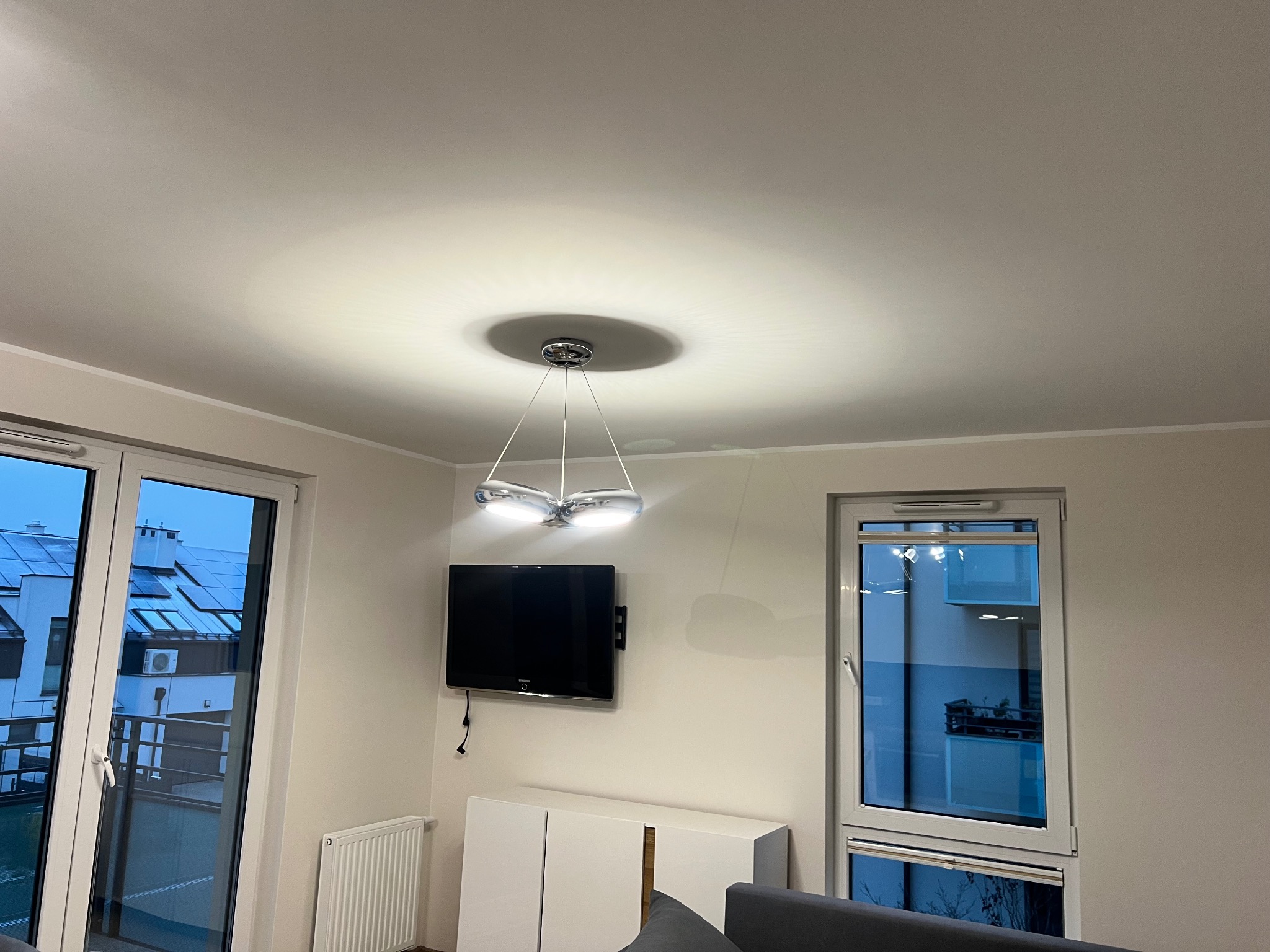 Jasne wnętrze pokoju z nowoczesną lampą sufitową, telewizorem na ścianie i białymi meblami. Widok na zewnątrz przez okno balkonowe.