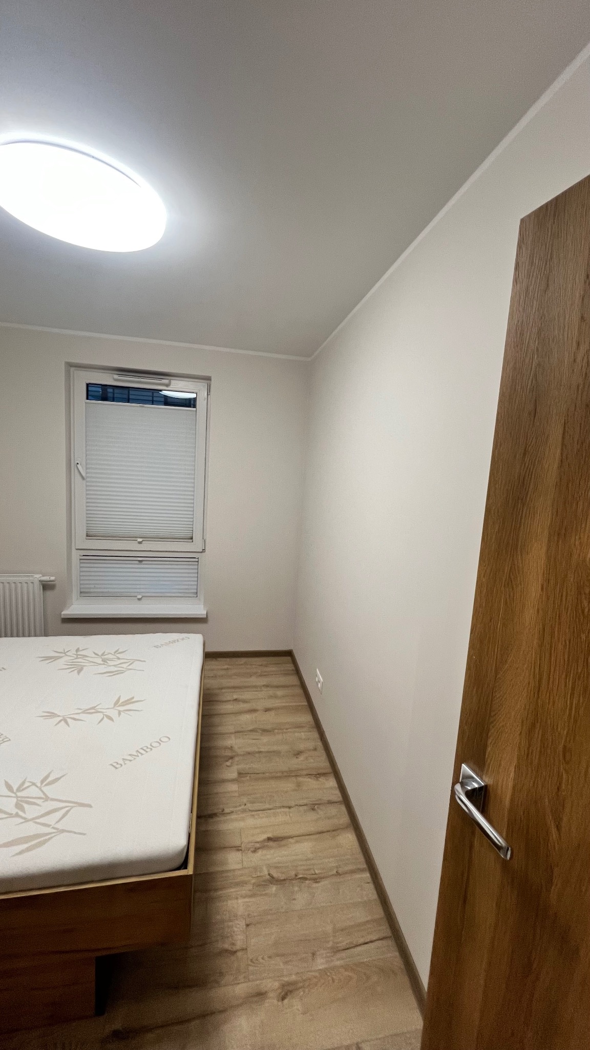 Jasne wnętrze sypialni po remoncie: panele podłogowe, pomalowane ściany w odcieniach bieli, okno z żaluzjami plisowanymi i drewniane drzwi z metalową klamką.