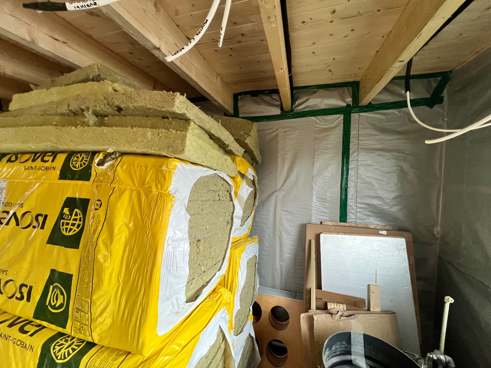 Paczki wełny mineralnej Isover na tle drewnianej konstrukcji sufitu i folii izolacyjnej. Widoczne elementy instalacji elektrycznej.