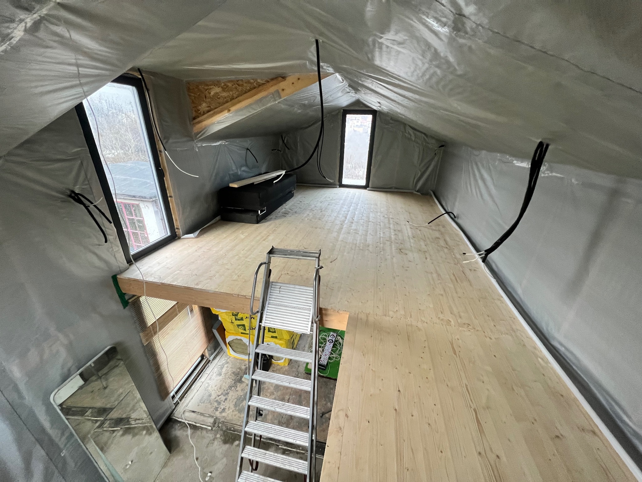 Poddasze w trakcie adaptacji: drewniana podłoga, okna dachowe, folia ochronna na ścianach i suficie, drabina. Widok na fragment niższej kondygnacji.