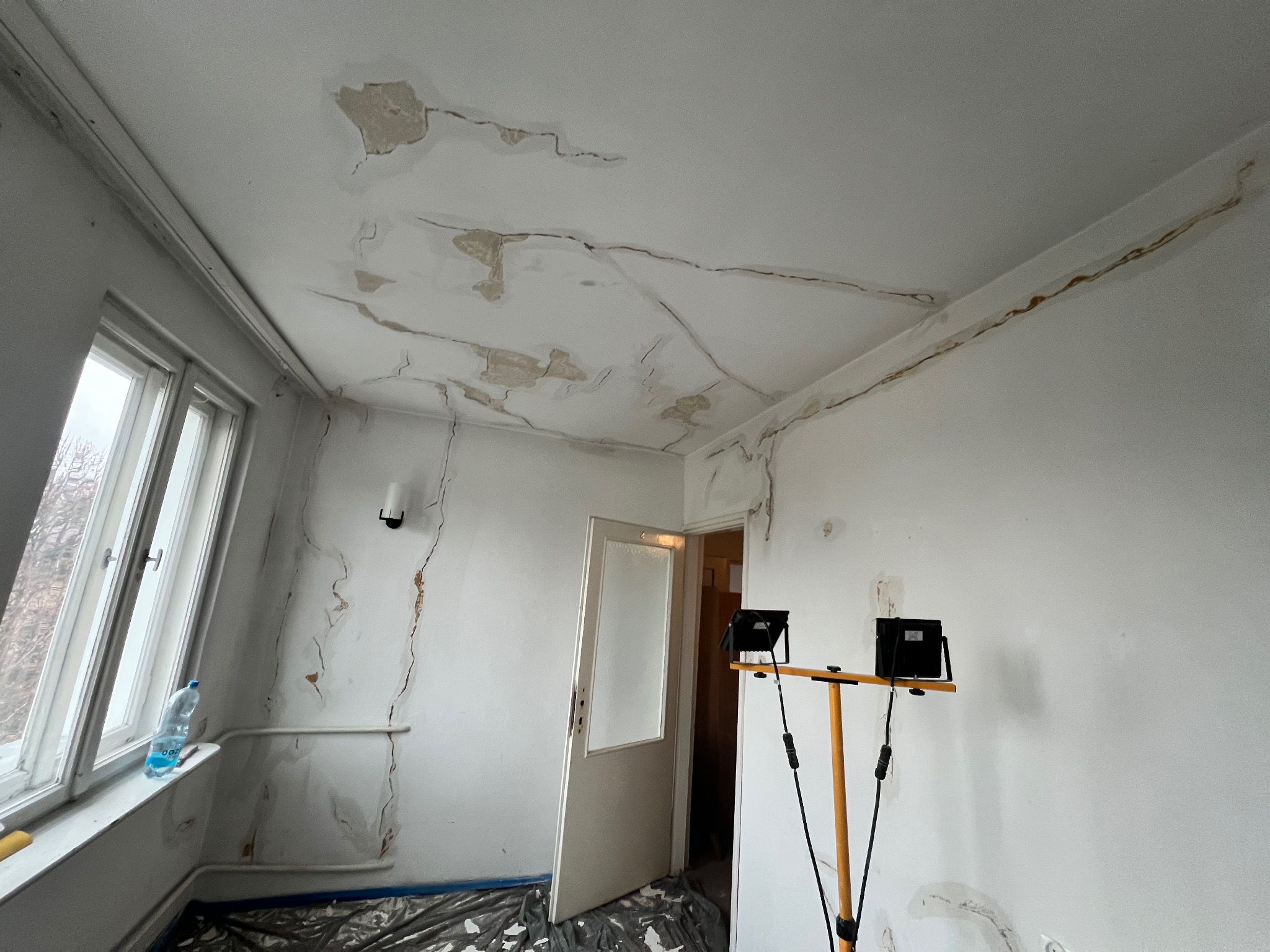 Pokój przed remontem: zacieki na suficie i ścianach, widoczne ślady wilgoci, zabezpieczona podłoga folią. Lampy oświetlają obszar wymagający naprawy.