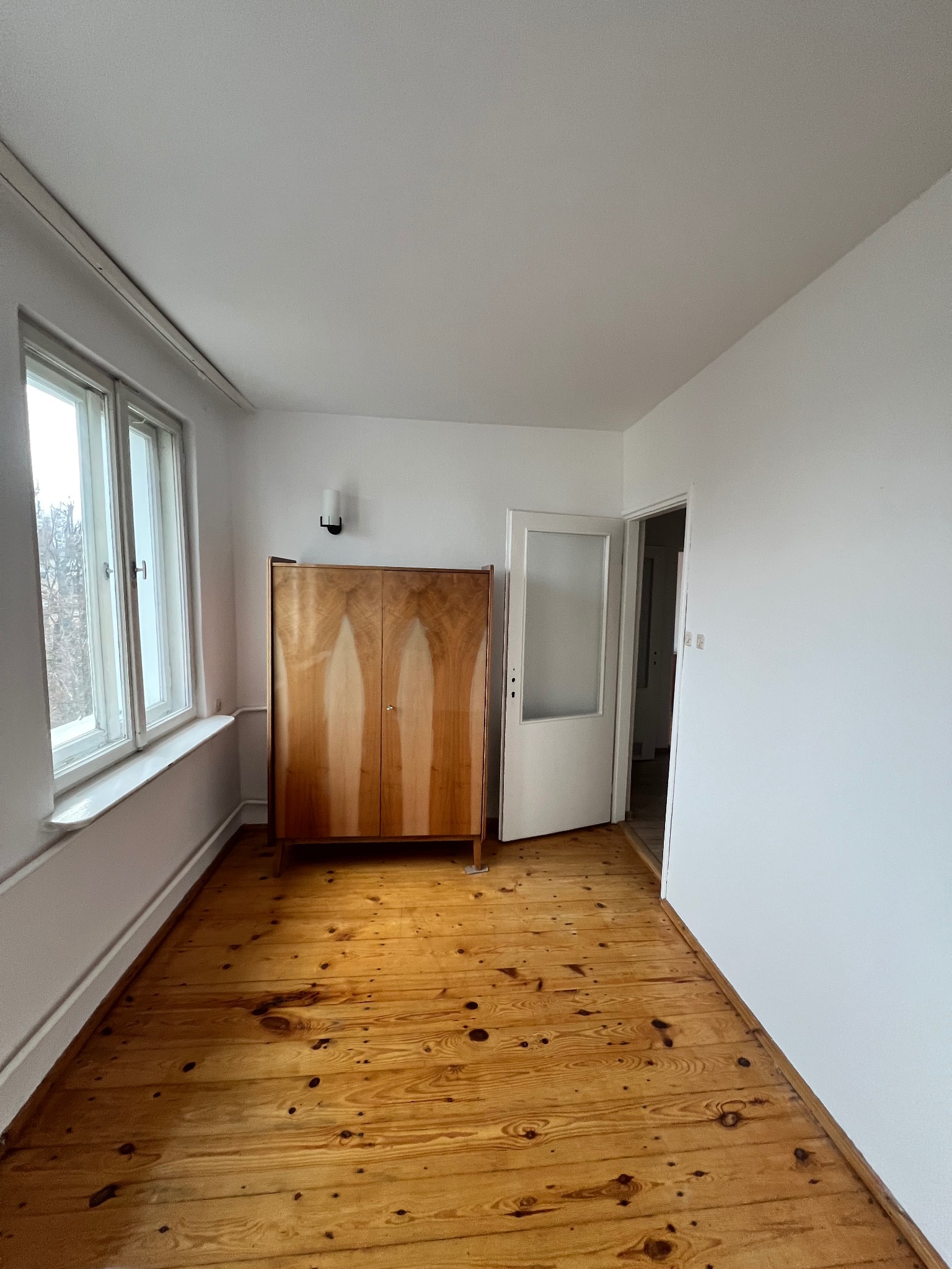 Jasne, puste wnętrze pokoju po remoncie: świeżo pomalowane ściany, odnowiona drewniana podłoga, okno z widokiem na zewnątrz i stylowa szafa w rogu.