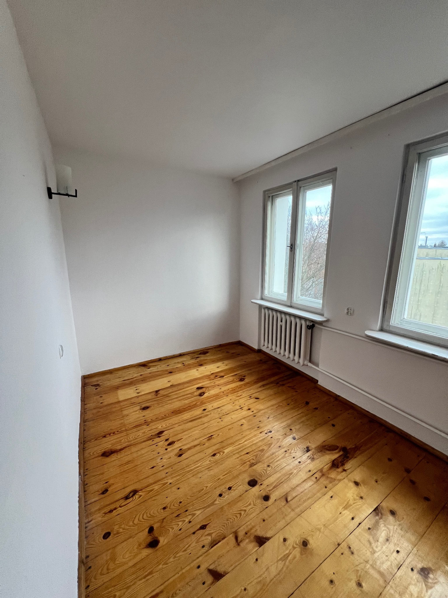 Pusty pokój z drewnianą podłogą, białymi ścianami i dwoma oknami. Widoczny grzejnik pod oknem. Minimalistyczne wnętrze po remoncie.