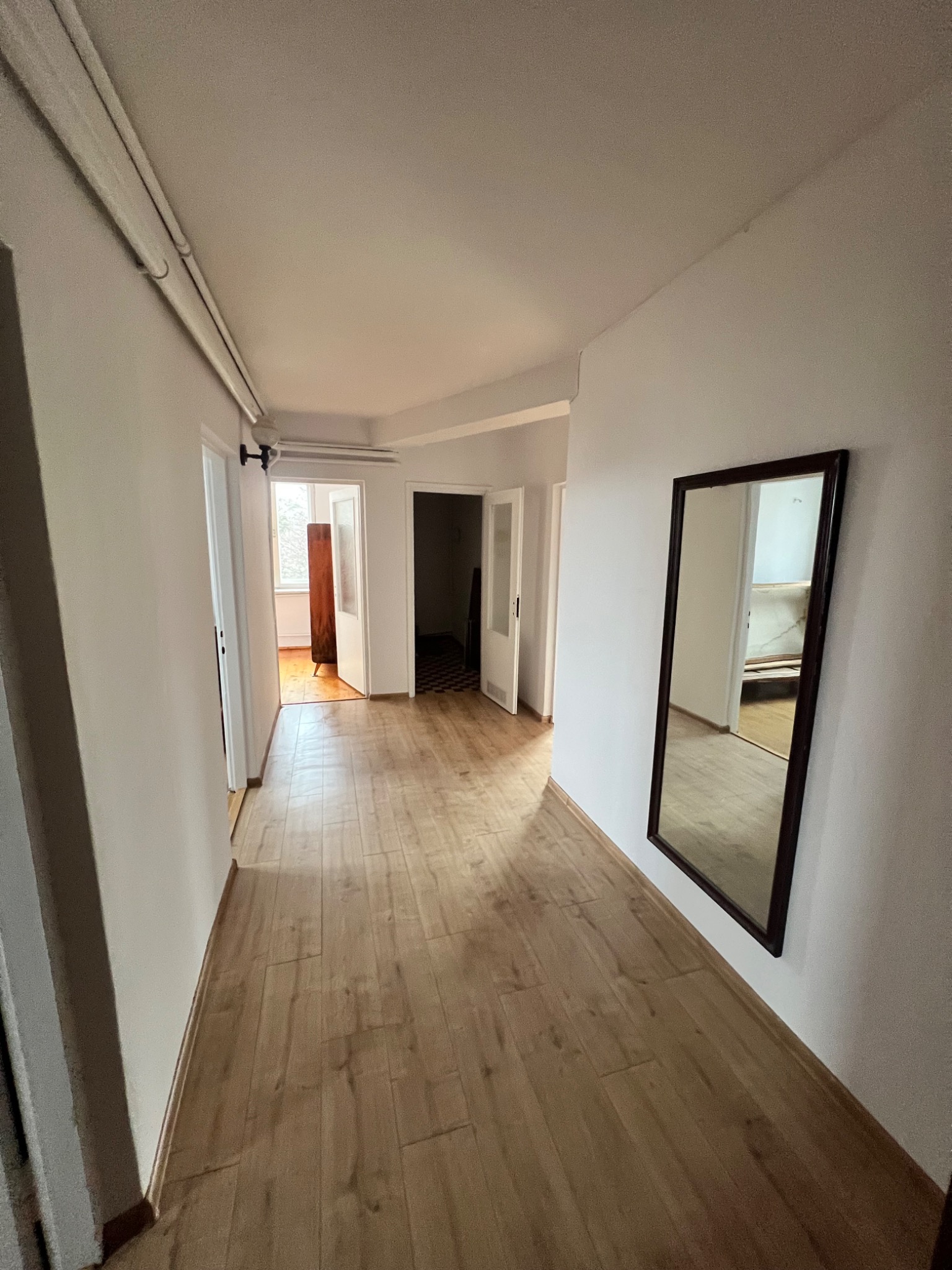 Jasny korytarz po remoncie z panelami podłogowymi i lustrem w ciemnej ramie. Widoczne drzwi do innych pomieszczeń i oświetlenie sufitowe.