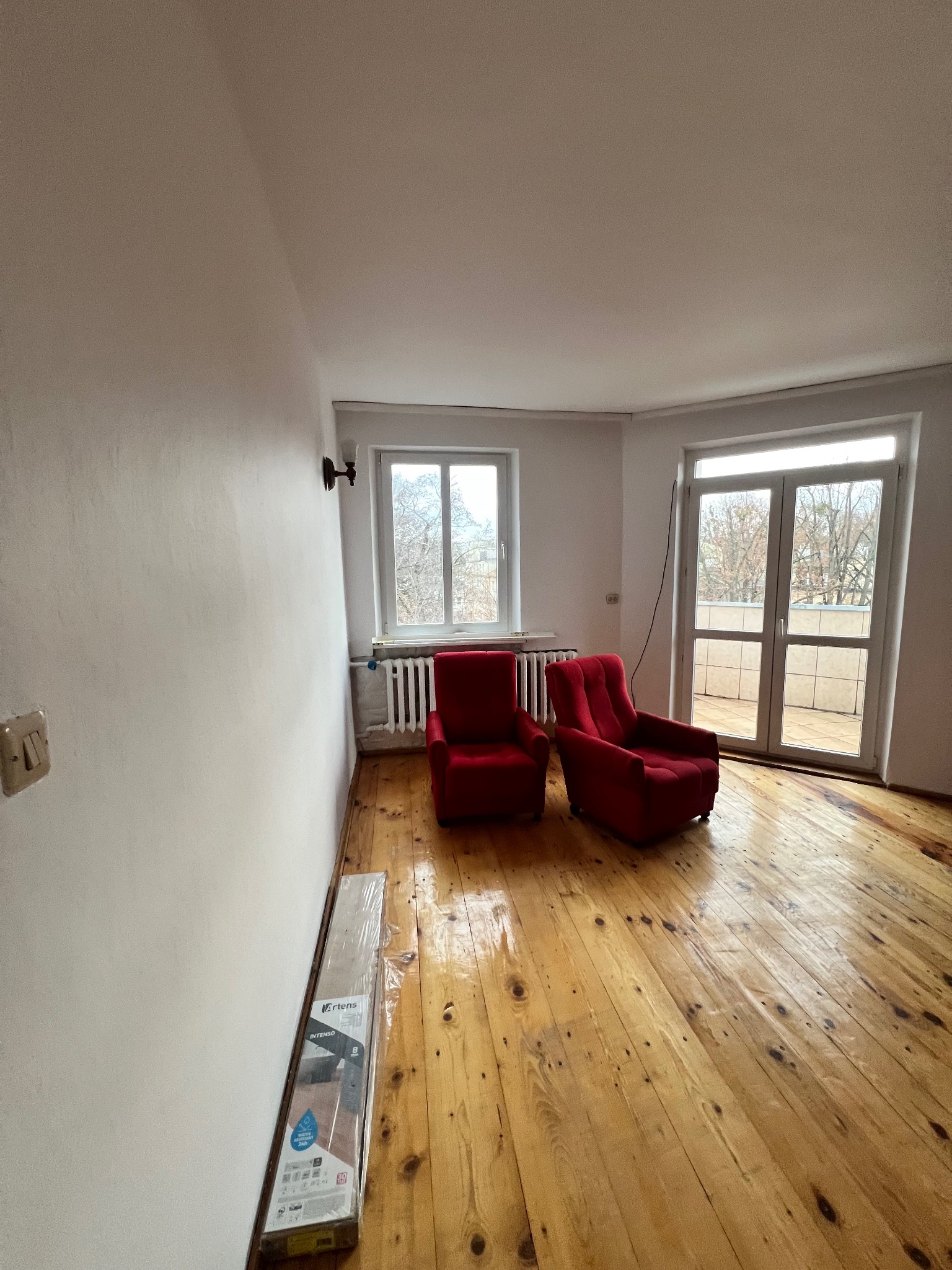 Jasne wnętrze z drewnianą podłogą, dwoma czerwonymi fotelami i oknami. Na podłodze leży zapakowany parkiet, gotowy do ułożenia. Ściany białe, przygotowane do malowania.