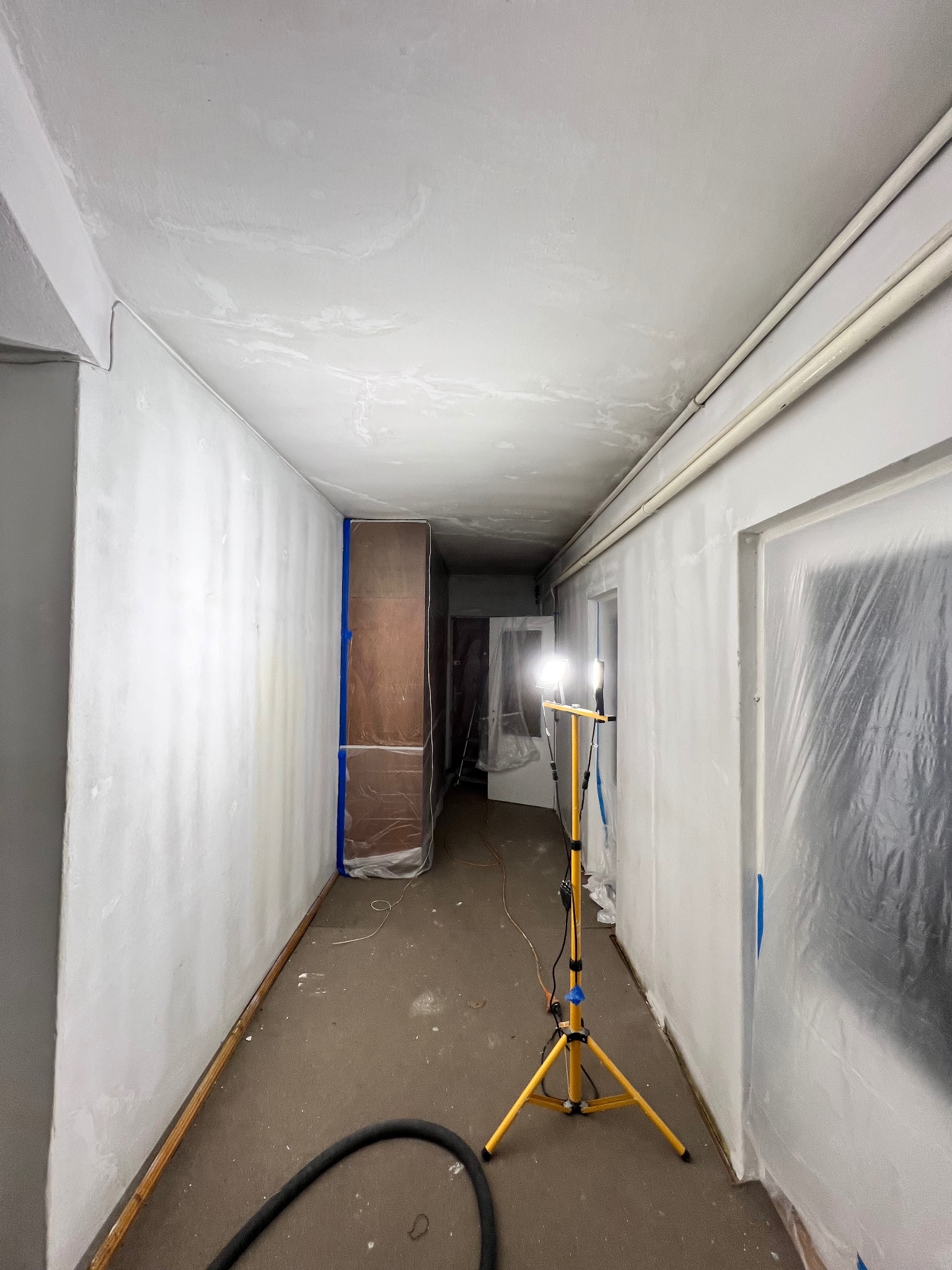Korytarz w trakcie przygotowań do malowania: ściany zagruntowane, meble i drzwi zabezpieczone folią, lampa budowlana oświetla przestrzeń. Widoczny wąż odkurzacza.