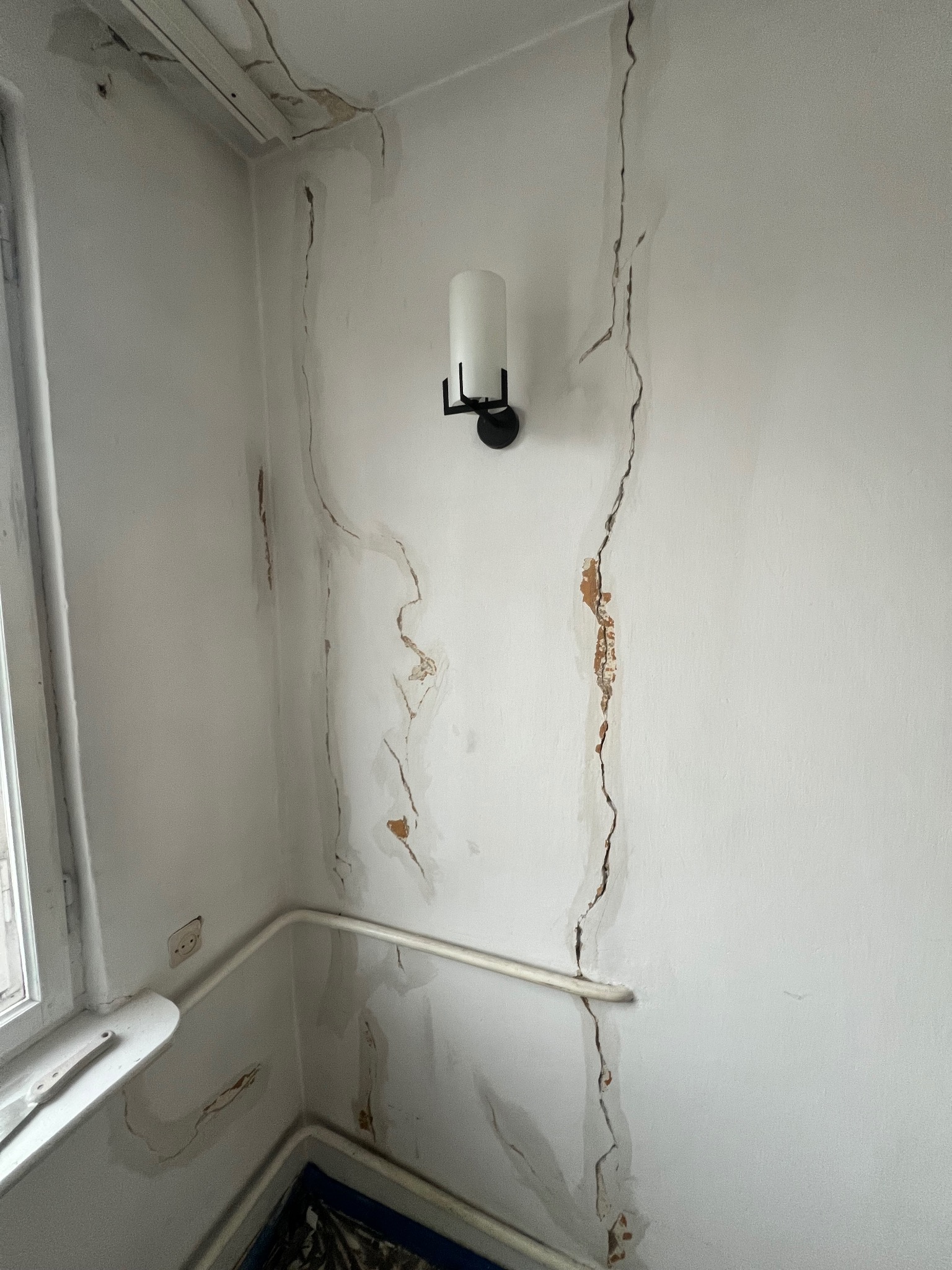 Pęknięcia i ślady wilgoci na ścianie w rogu pokoju, widoczna lampa ścienna i fragment okna. Ściana przygotowana do remontu, widoczne uszkodzenia tynku.