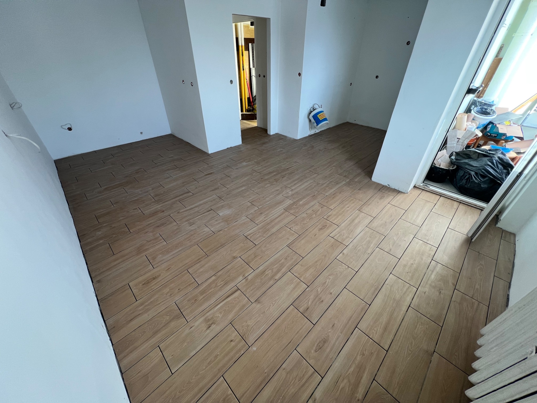 Ułożona podłoga z paneli imitujących drewno w pustym, jasnym pomieszczeniu z białymi ścianami. Widoczne gniazdka elektryczne i otwarte drzwi.