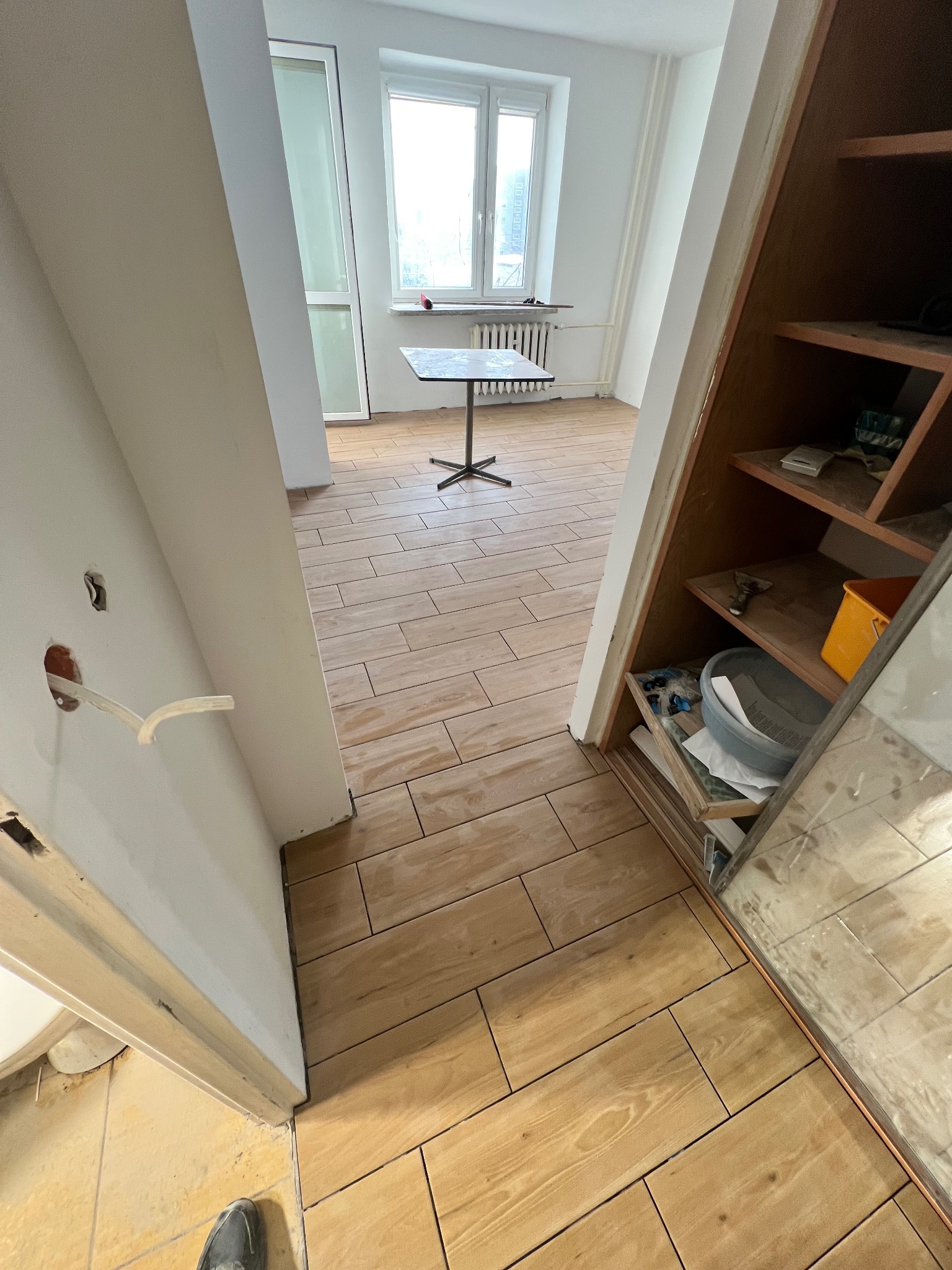 Ułożona podłoga z płytek imitujących drewno w jasnym pokoju, widok z korytarza. Otwarta szafa z narzędziami po prawej, okno w tle.