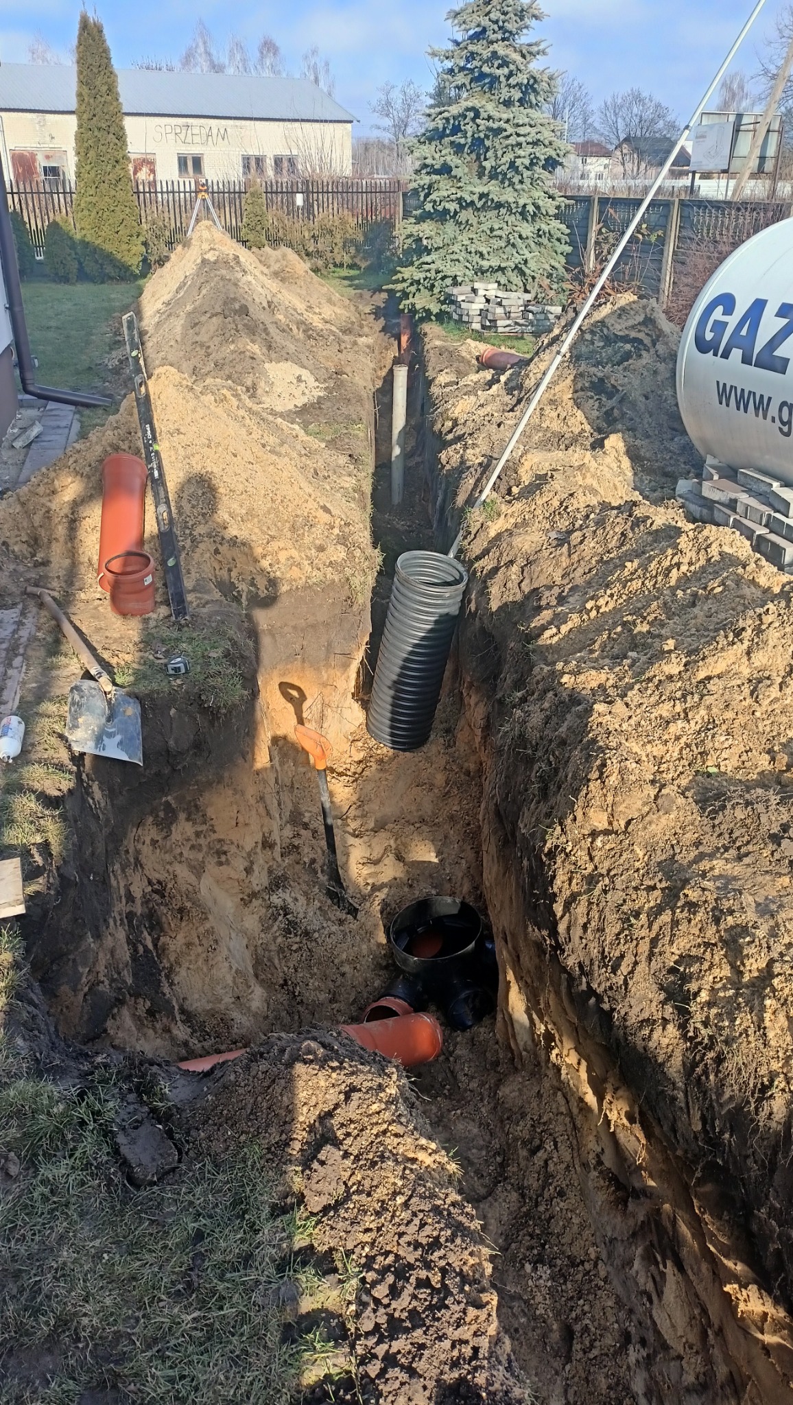 Głęboki wykop w ziemi z widocznymi rurami kanalizacyjnymi, szpadlem i poziomicą. W tle budynek z napisem 'Sprzedam' i zbiornik gazu.