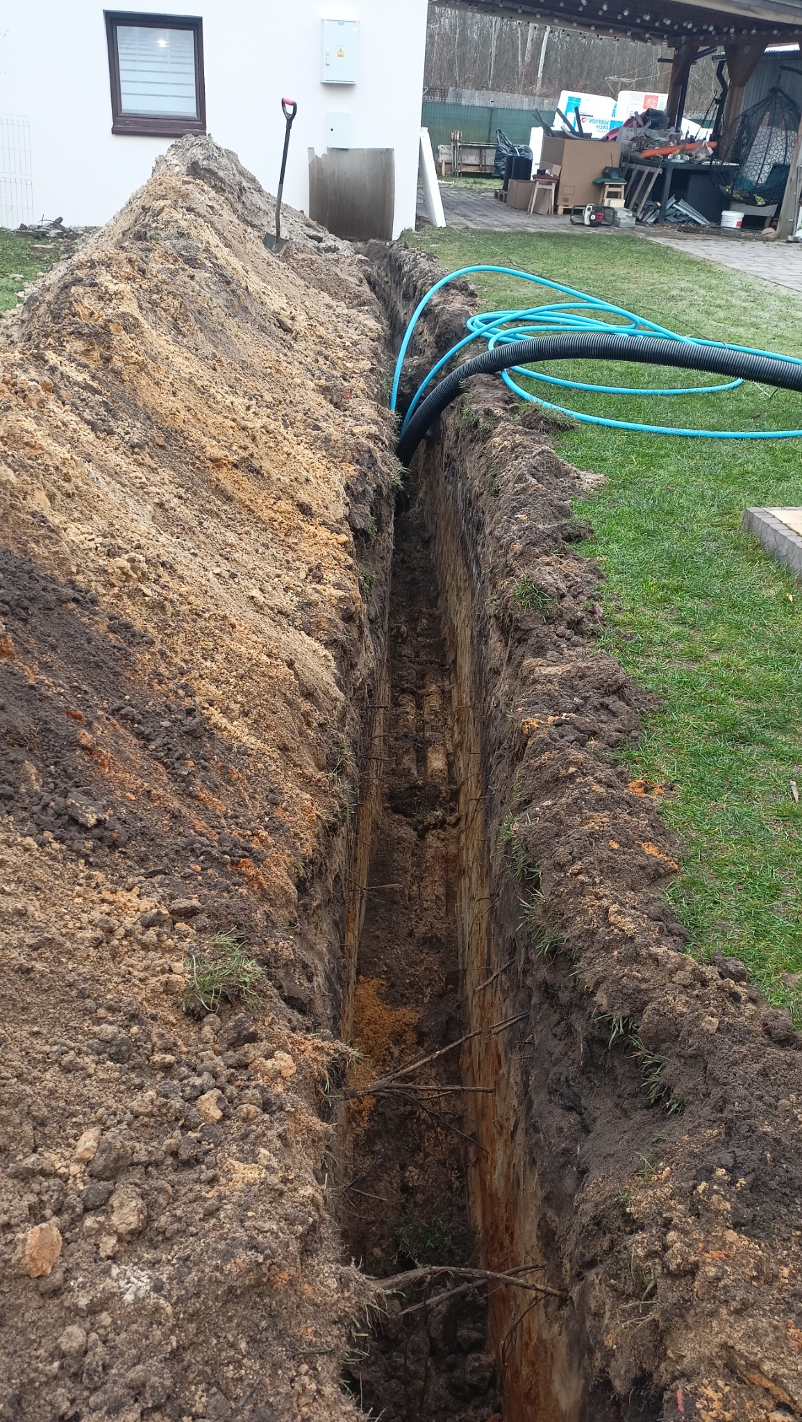 Wąski wykop w ziemi z ułożonymi niebieskimi rurami, obok sterta ziemi i łopata. Widoczny budynek w tle. Prace ziemne w trakcie realizacji.
