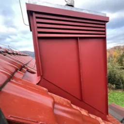 Czerwony komin z metalową obudową i ozdobnym wiatrowskazem na błyszczącym, ceramicznym dachu. W tle jesienny krajobraz i pochmurne niebo.