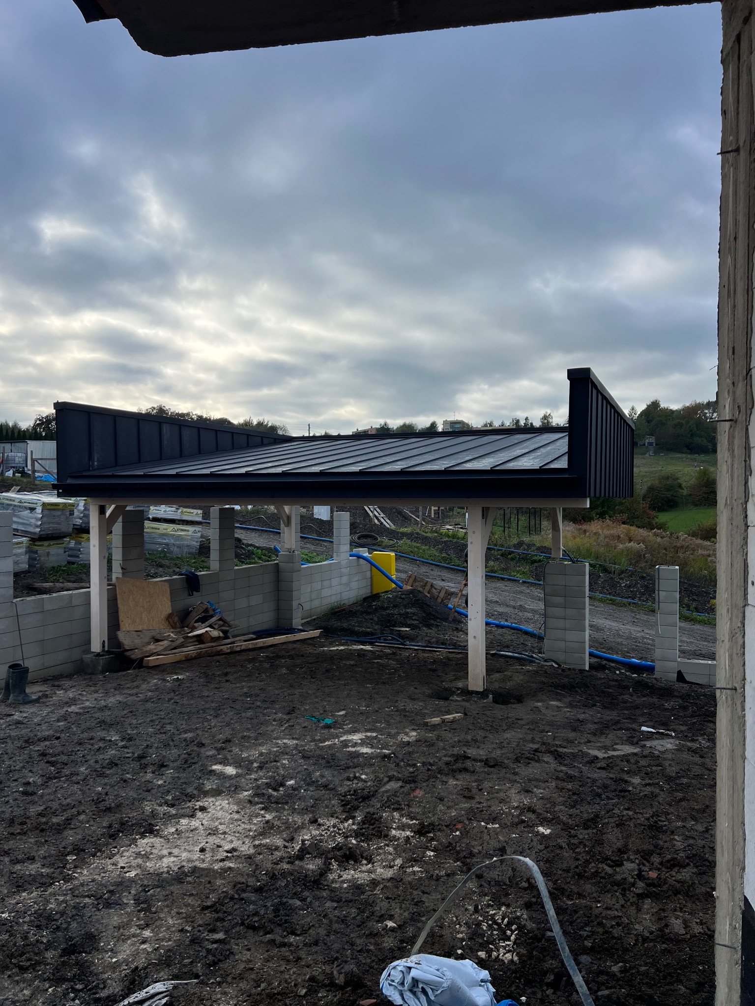 Nowoczesna, czarna konstrukcja dachu z blachy na drewnianych podporach w trakcie budowy. Widok z wnętrza budynku na zewnątrz, pochmurne niebo w tle.