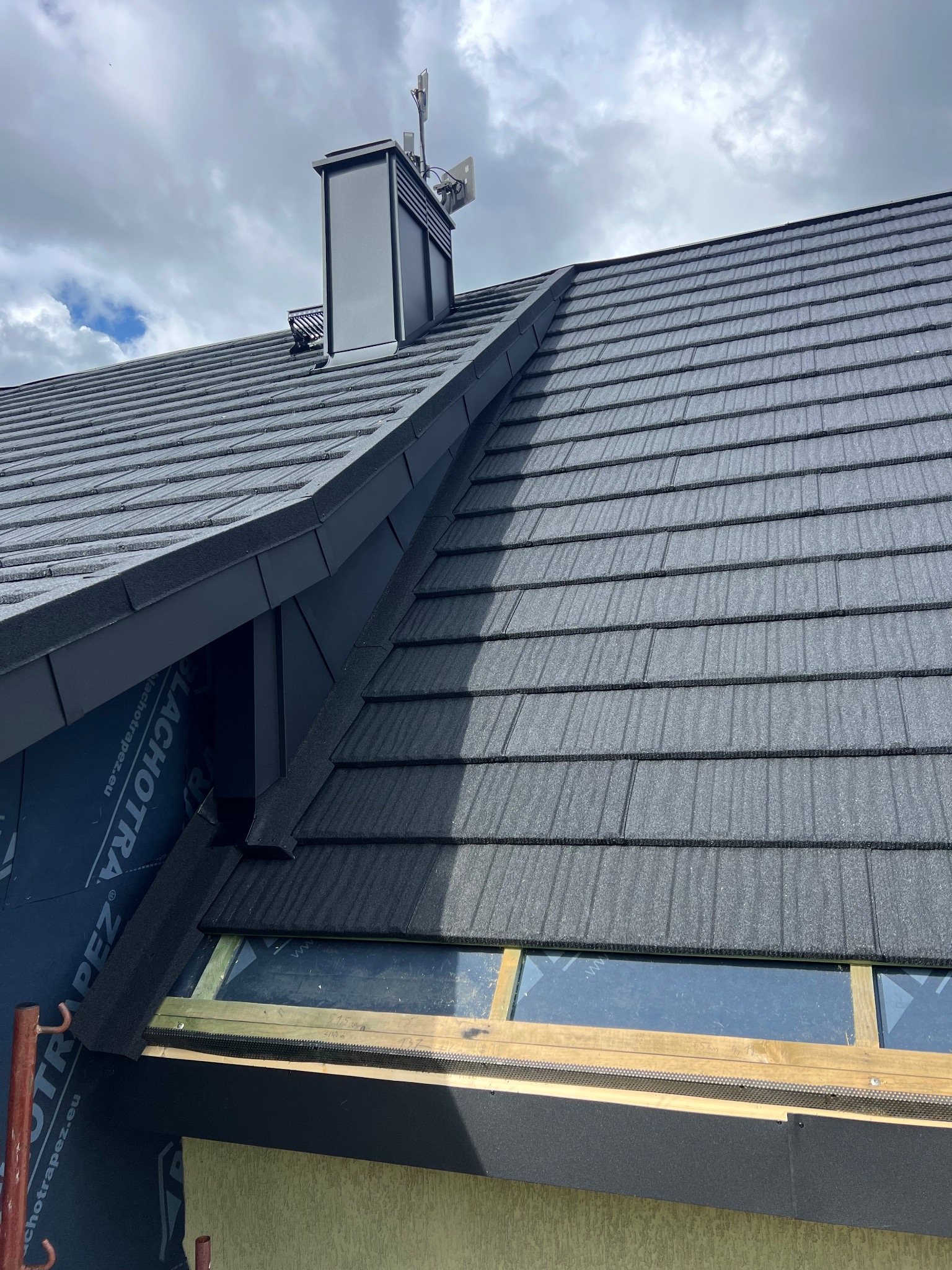 Fragment dachu z czarną dachówką, kominem i antenami na tle pochmurnego nieba. Widoczne detale obróbek blacharskich i okno dachowe w trakcie montażu.