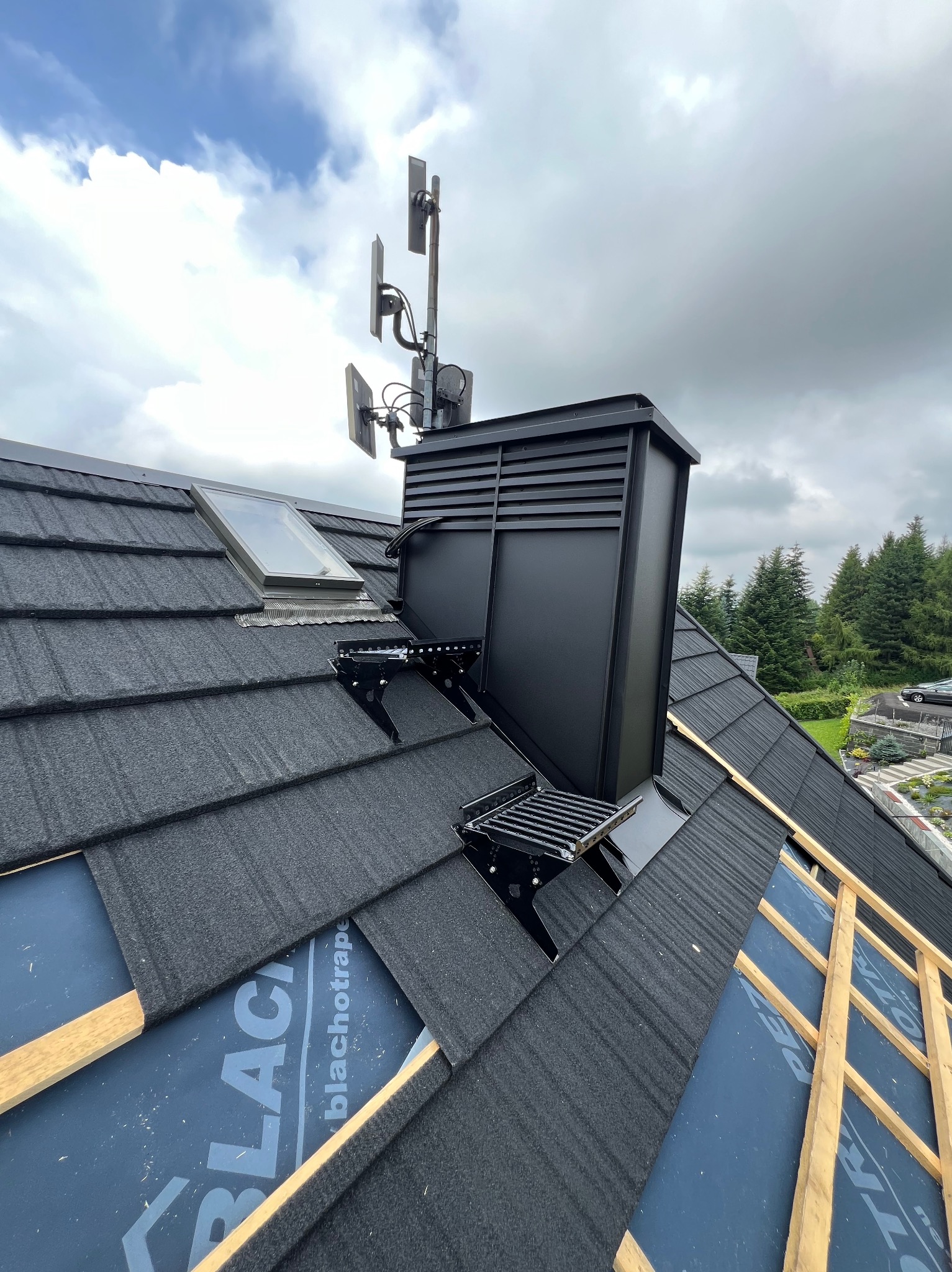 Czarny dach z blachodachówki modułowej, komin wentylacyjny z anteną. Widoczne okno dachowe i fragment więźby. W tle zieleń drzew i pochmurne niebo.