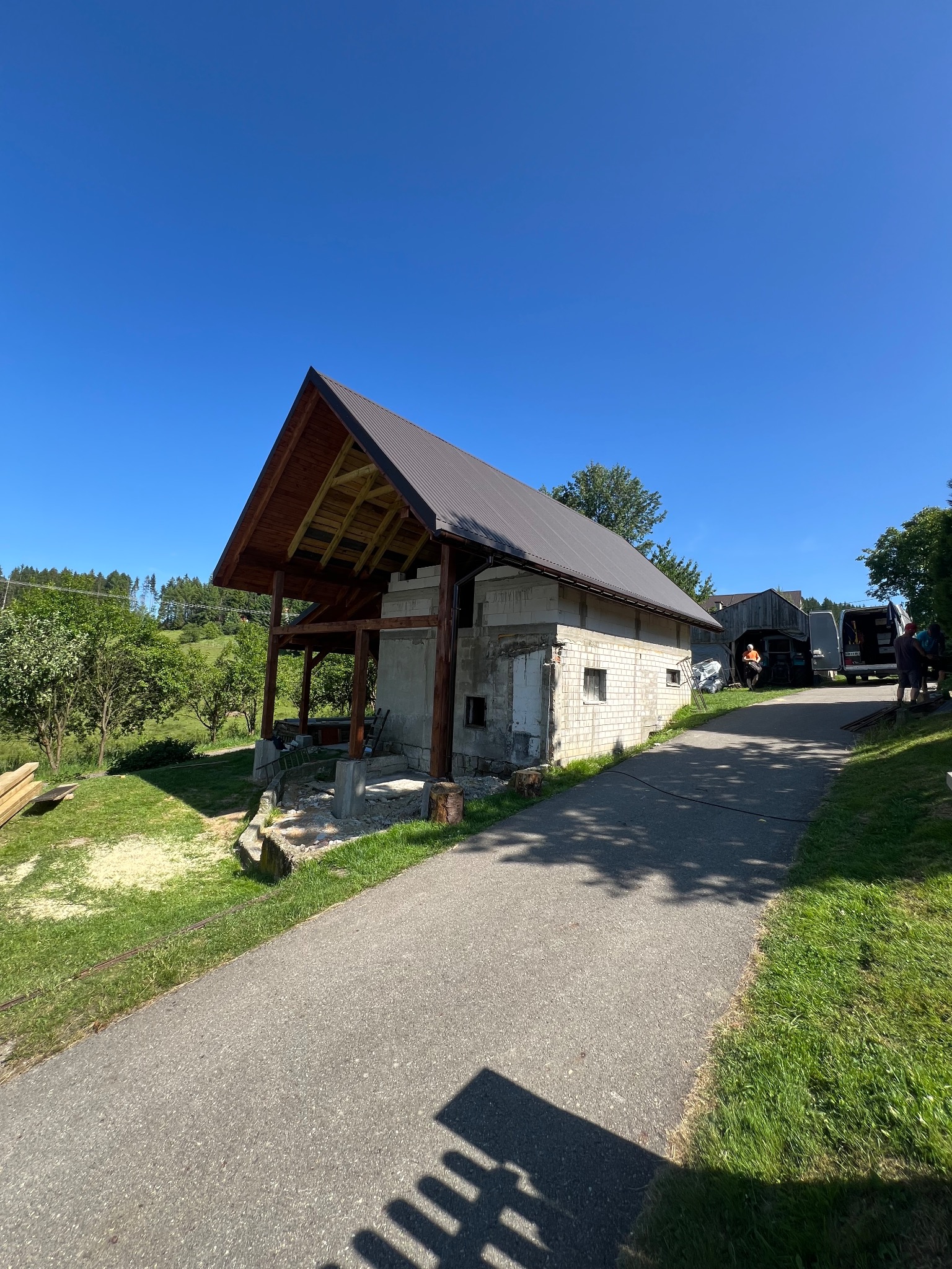 Budynek w trakcie budowy dachu z widoczną więźbą i częściowo pokrytym dachem z blachy na tle błękitnego nieba i zieleni. Widoczne materiały budowlane.