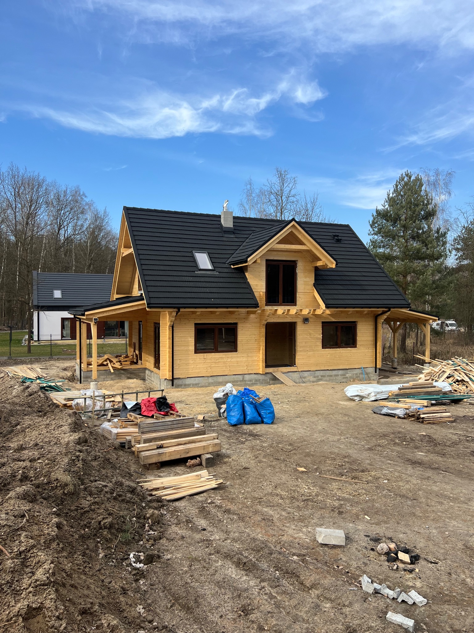 Surowy drewniany dom z czarnym dachem z dachówki, w trakcie budowy, z widoczną konstrukcją i materiałami budowlanymi na placu budowy. Błękitne niebo w tle.