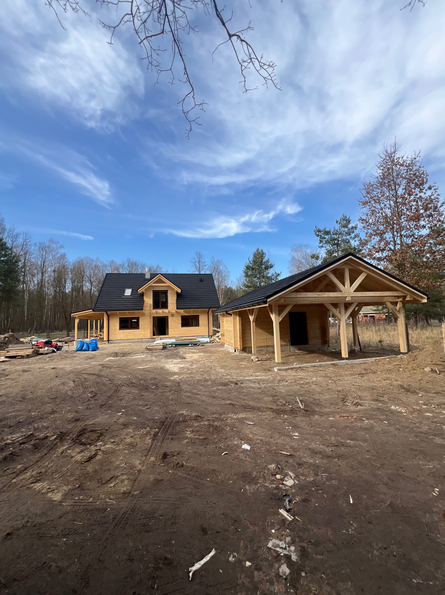 Surowy drewniany dom z czarnym dachem i drewniana wiata na tle błękitnego nieba, w trakcie budowy, z widocznymi materiałami budowlanymi.