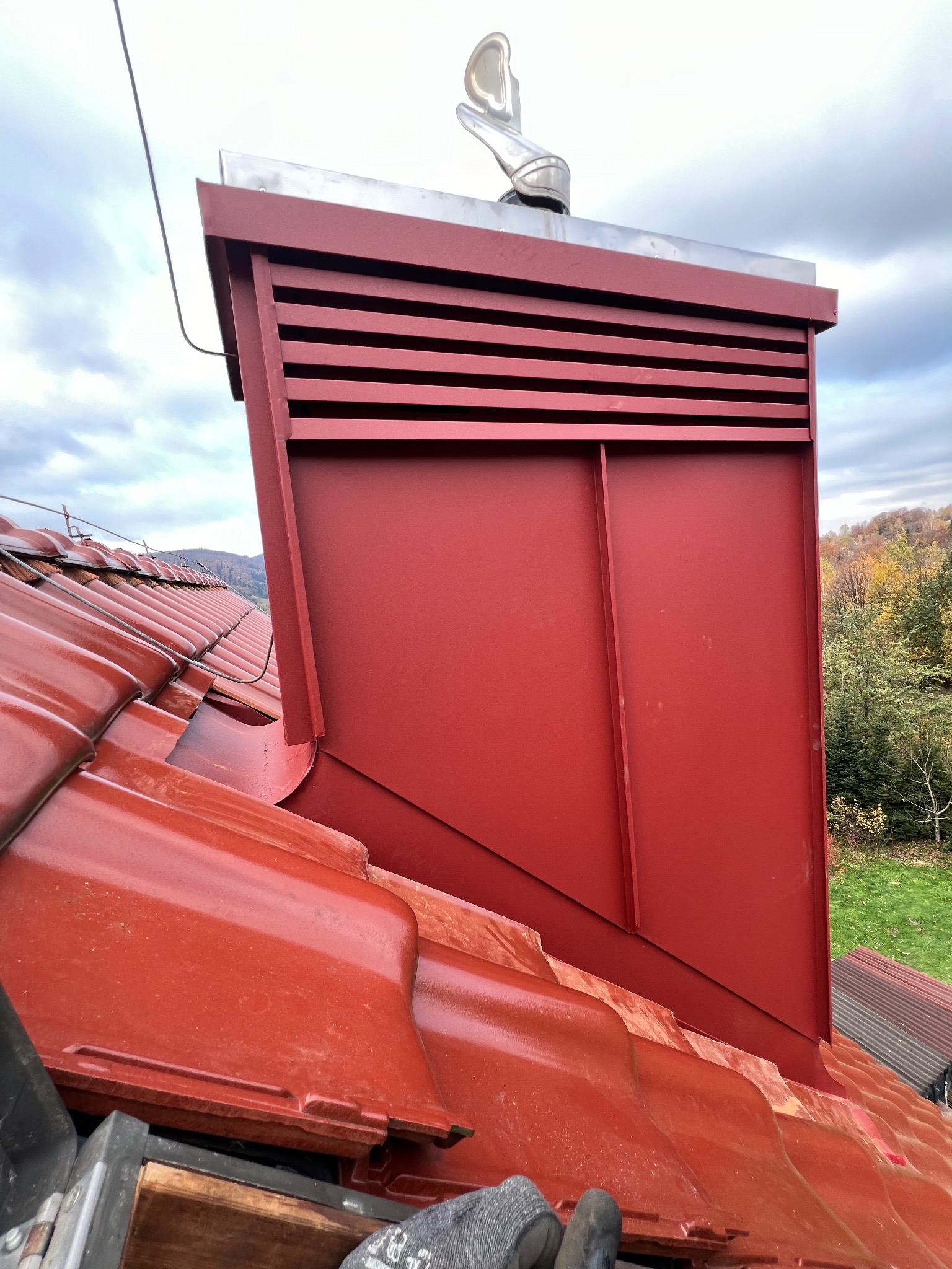 Czerwony komin z metalową obudową i ozdobnym wiatrowskazem na błyszczącym, ceramicznym dachu. W tle jesienny krajobraz i pochmurne niebo.