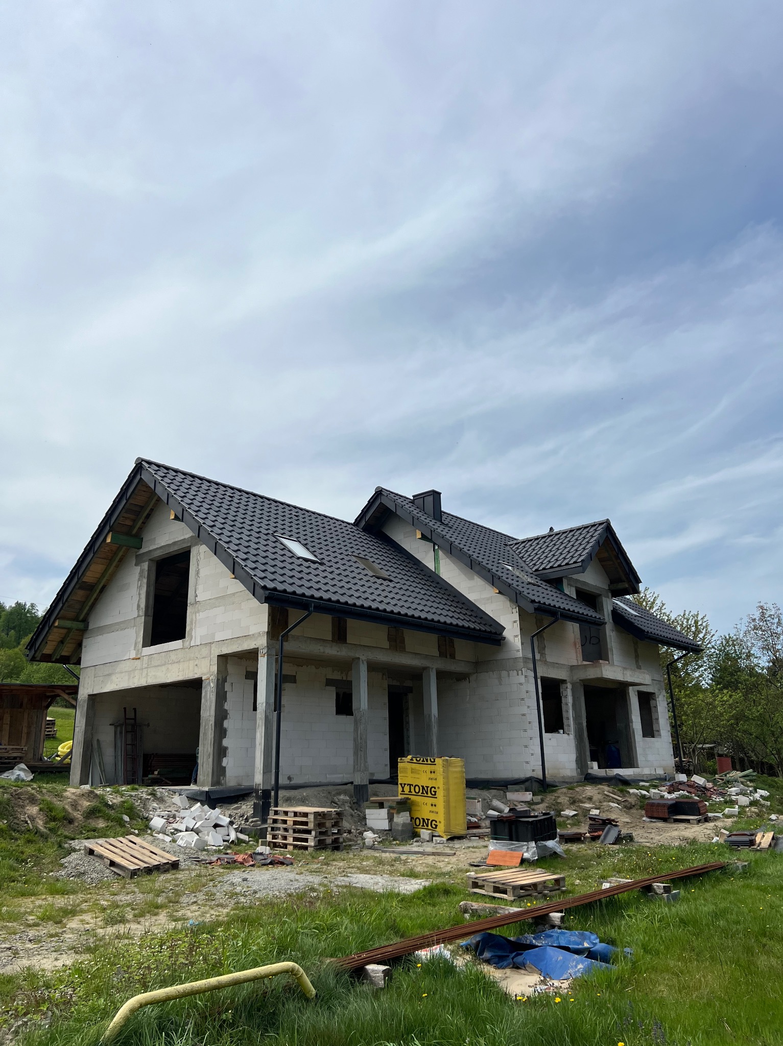 Dom w trakcie budowy z czarnym dachem z dachówki ceramicznej. Widoczne bloczki YTONG, drewniane palety i zbrojenia. Budynek w stanie surowym zamkniętym.
