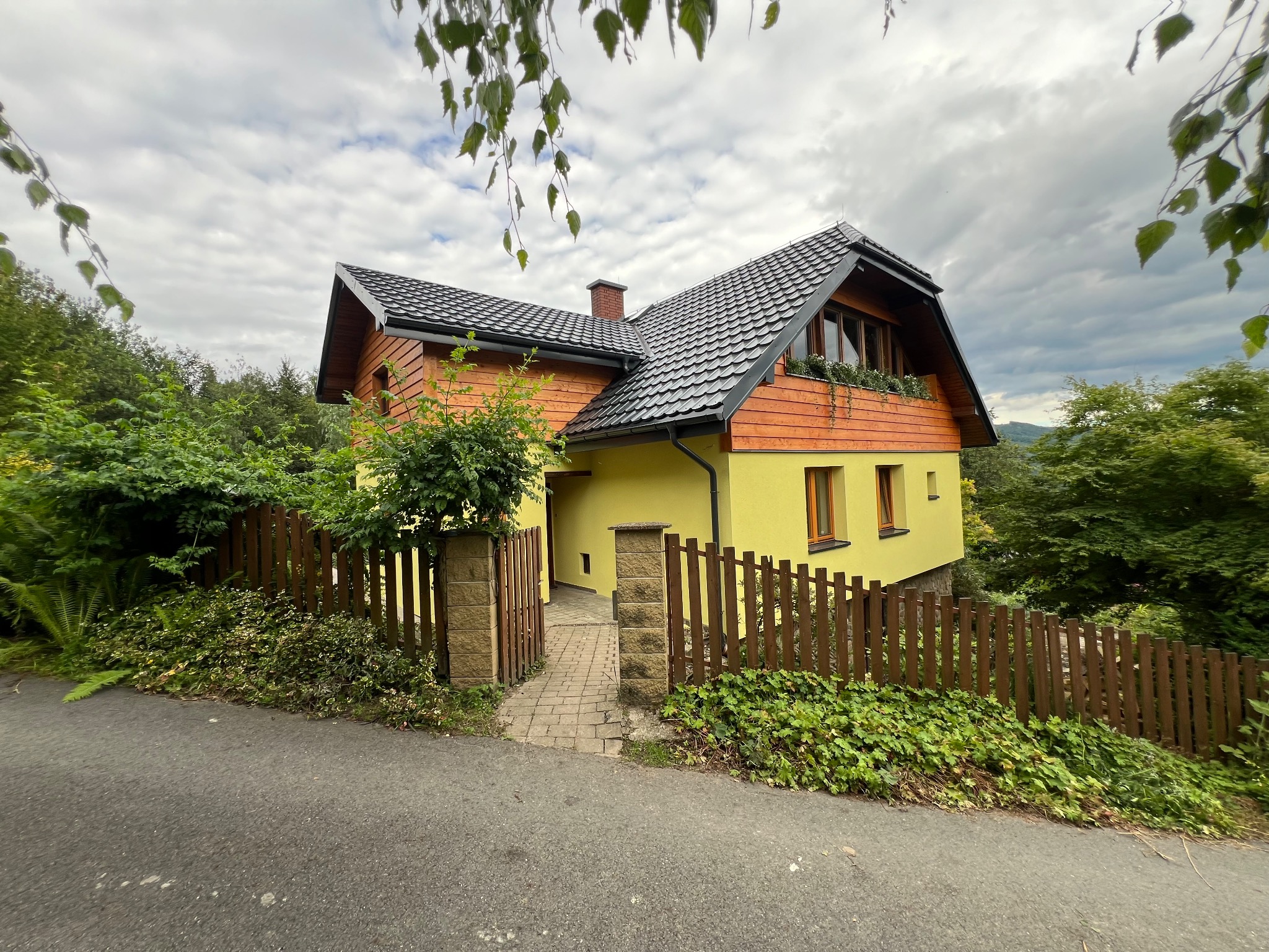 Żółty dom z ciemnym dachem i drewnianą elewacją, otoczony zielenią i niskim płotem. Widok z poziomu ulicy, pochmurne niebo w tle.